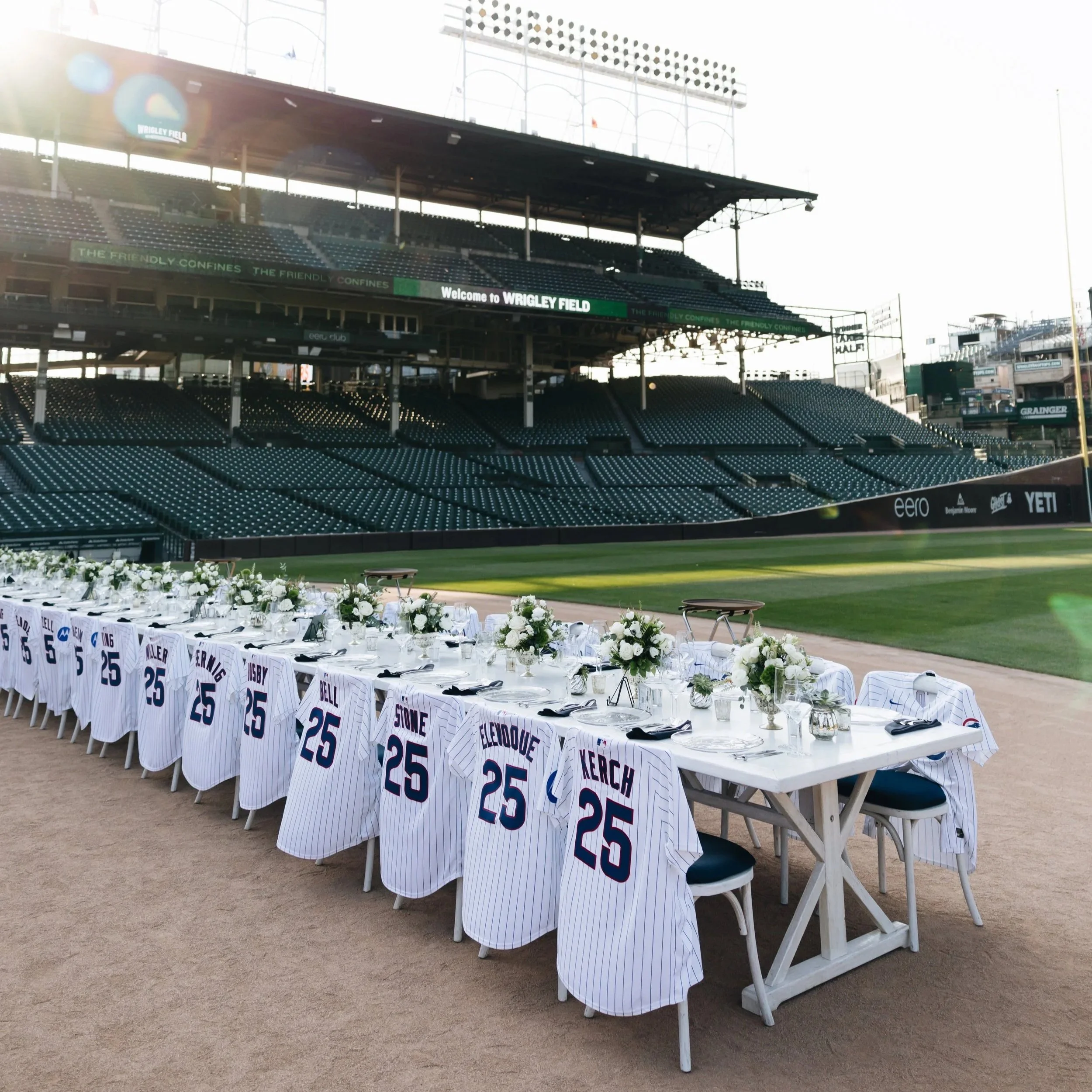 Empty baseball stadium with a long table set up with white chairs, white tablecloths, floral centerpieces, and sports jerseys hanging on the chairs, located on the field in front of the stands at Wrigley Field.