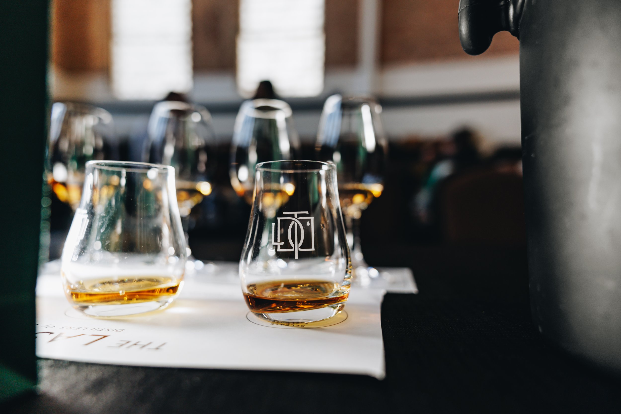 Close-up of three glasses of whiskey on a bar table, with additional glasses blurred in the background, taken through a dark opening.