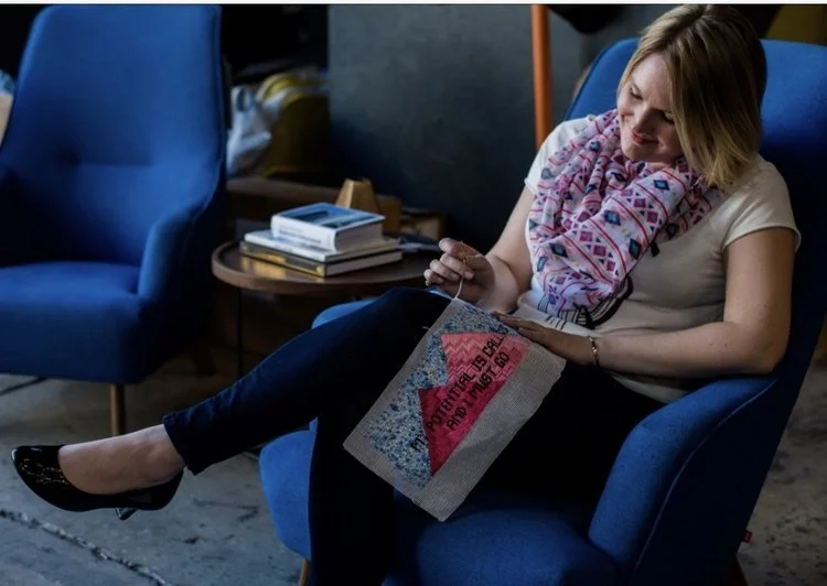 Kate sits in blue chair working on cross stitch