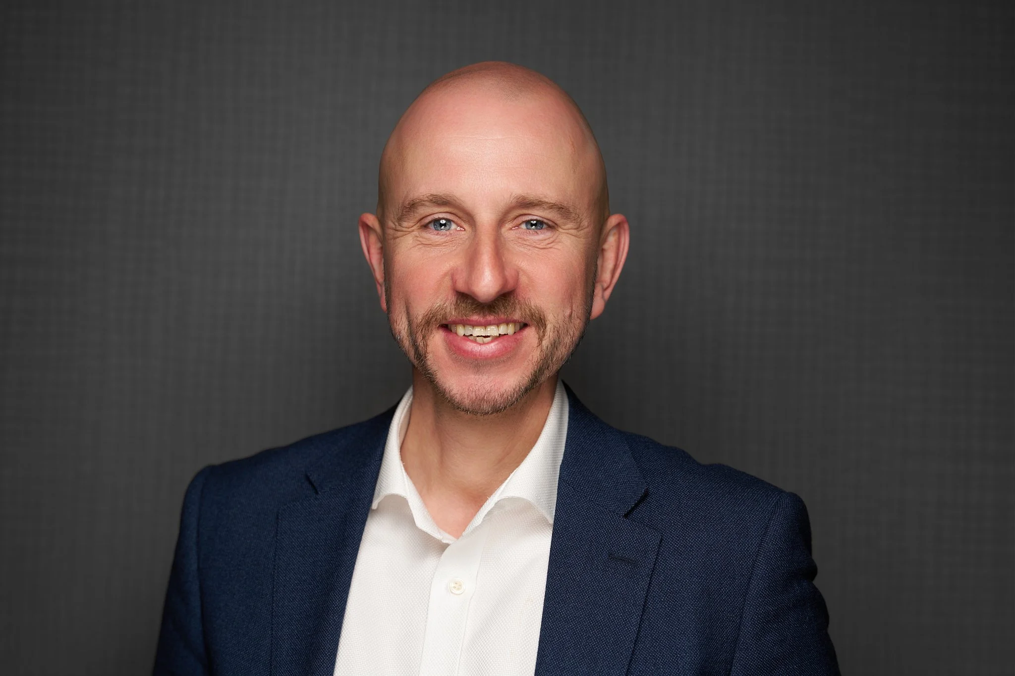 BESA staff headshot of a smiling bald man with a short beard wearing a navy blazer and white shirt against a dark grey background.