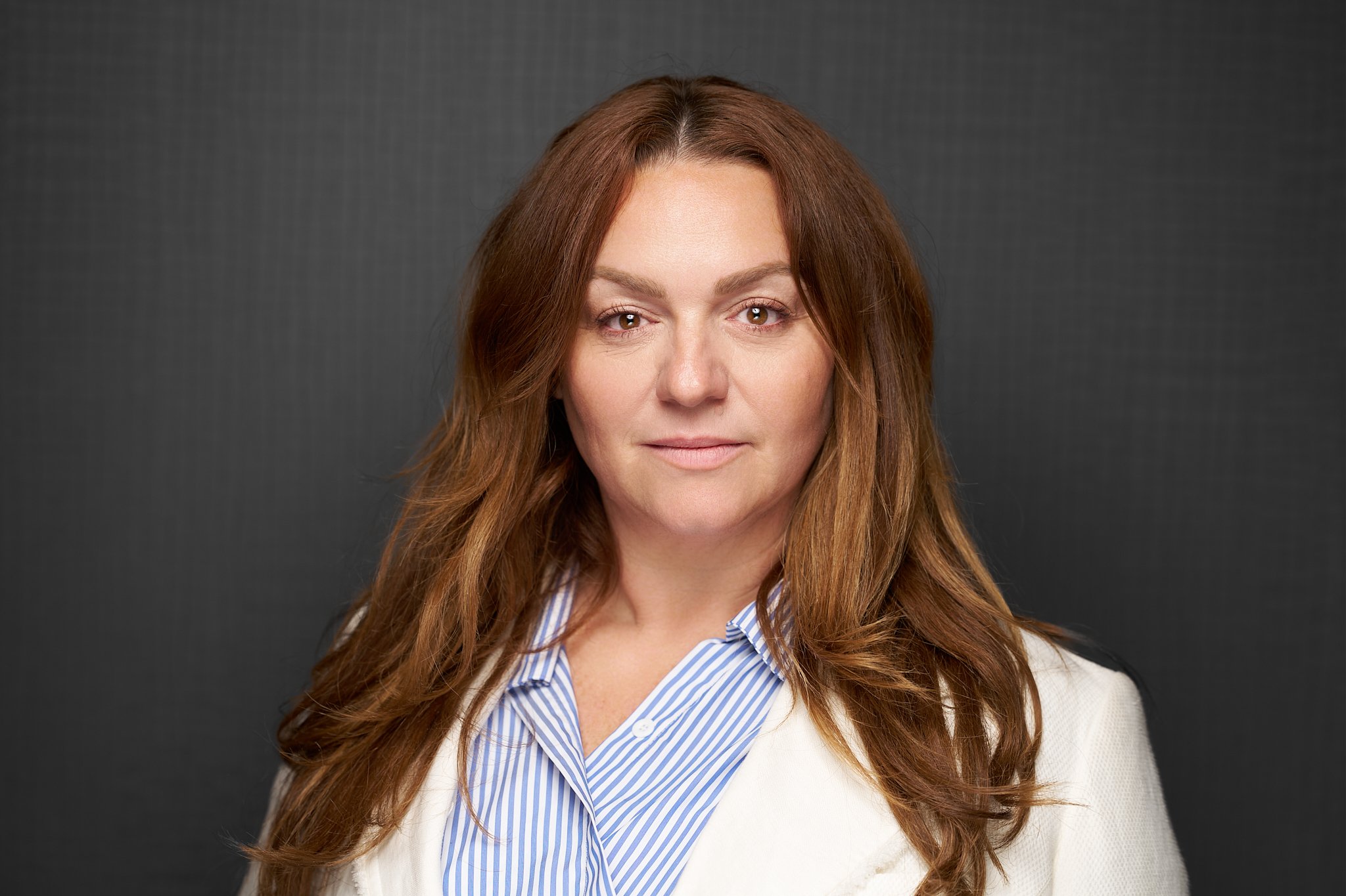 BESA staff headshot of a woman with long brown hair wearing a cream blazer and blue striped shirt against a dark grey background.