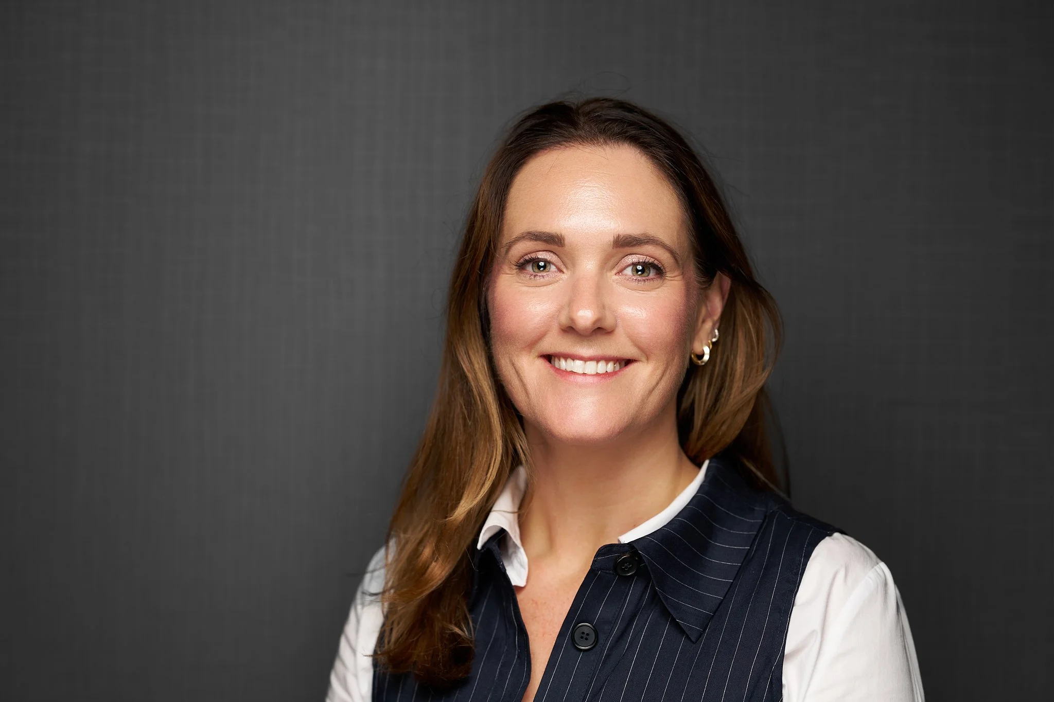 BESA staff headshot of a smiling woman wearing a navy pinstriped waistcoat over a white shirt against a dark grey background.