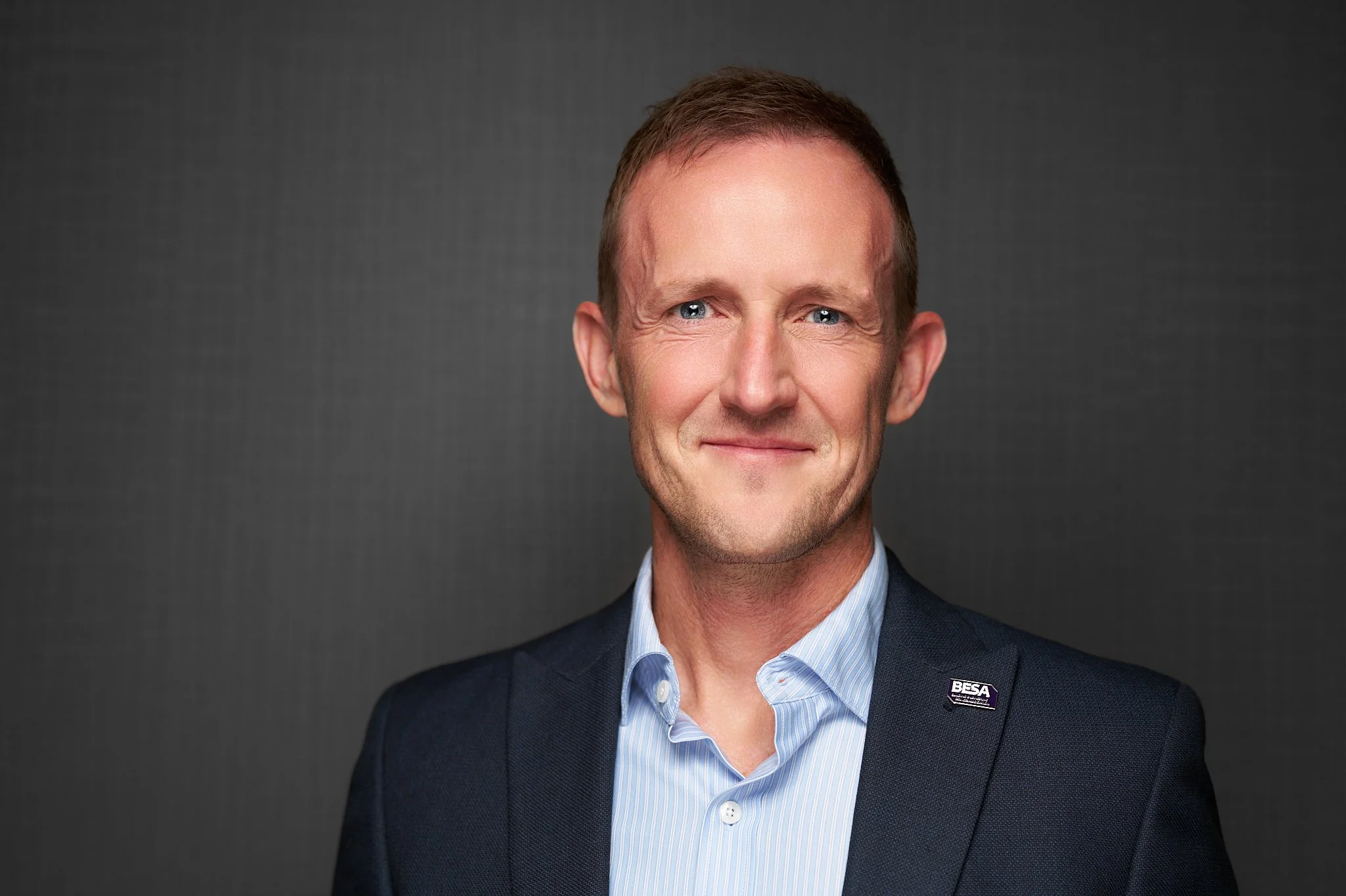 BESA staff headshot of a man wearing a navy blazer, light blue striped shirt and BESA lapel badge against a dark grey background.