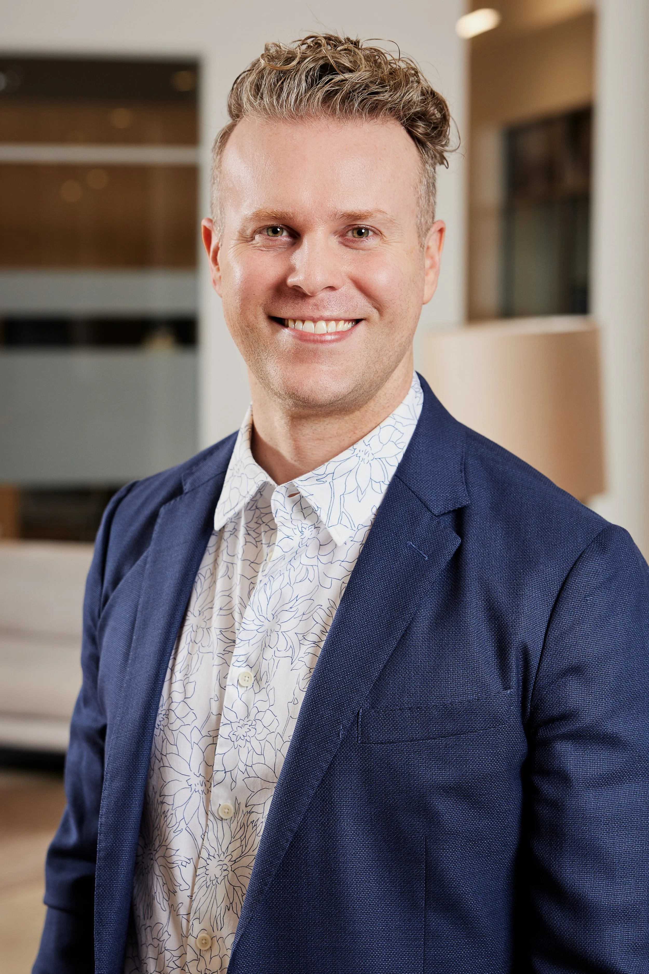 A smiling man, Patrick Shaw, with curly hair wearing a navy blazer and a white floral shirt, standing in an interior space with modern decor.