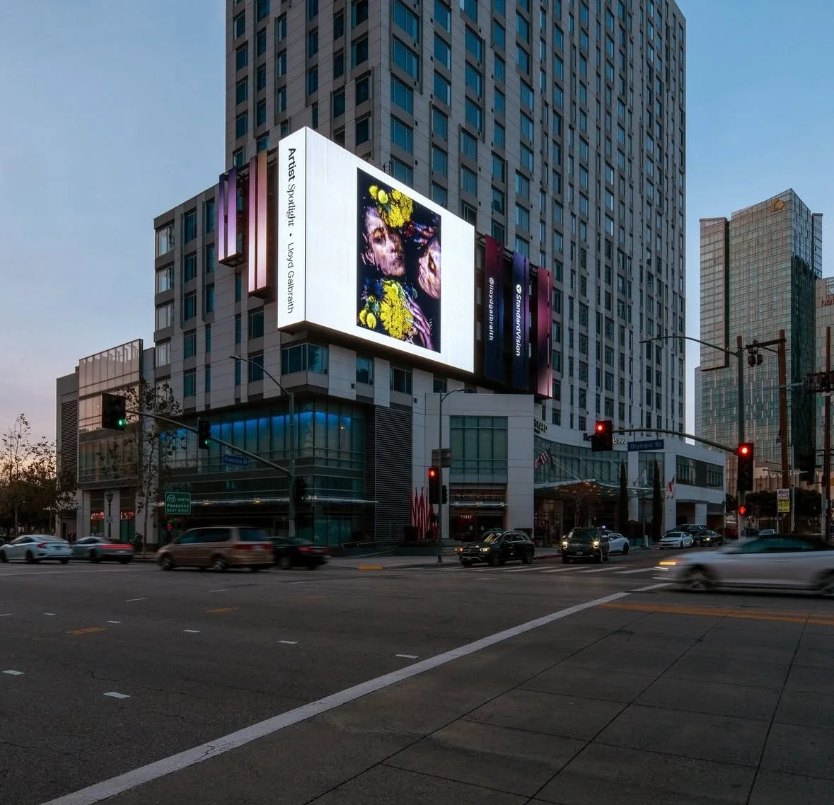 City street intersection with tall buildings, digital billboards, and cars passing by during dusk. One billboard shows abstract art of two faces with yellow flowers.