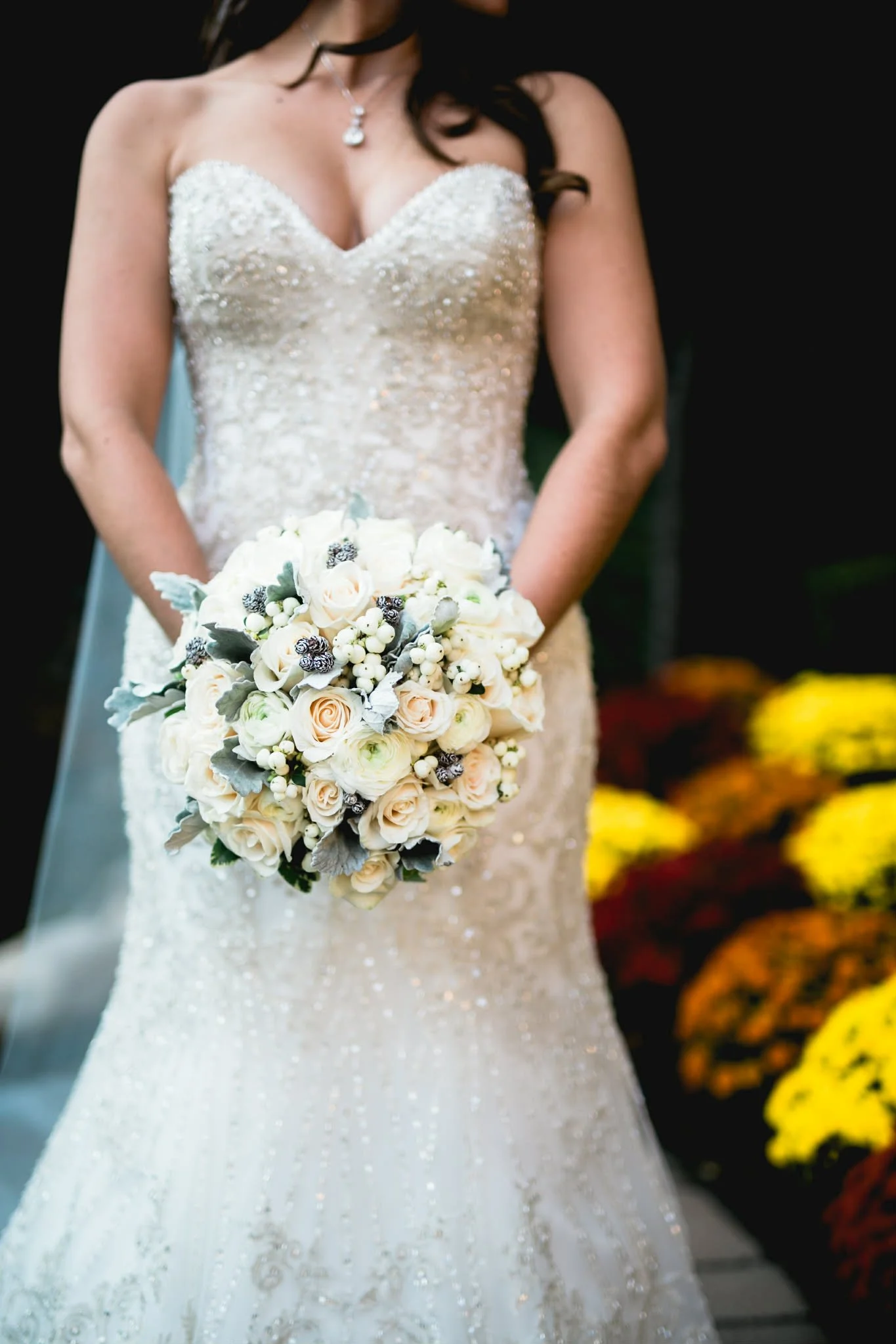 A bride in a white, beaded wedding gown holding a bouquet of white and light pink roses with silver accents, standing outdoors with colorful flowers in the background.