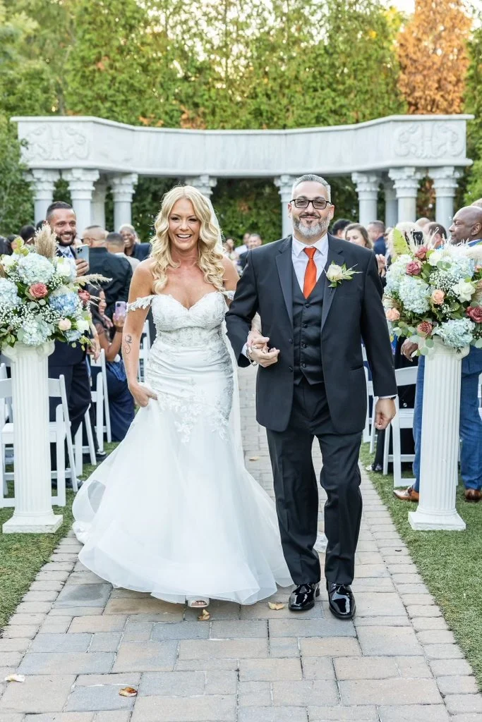 Bride and groom walking down the aisle at their outdoor wedding ceremony, surrounded by guests and floral arrangements, with greenery and a decorative structure in the background.
