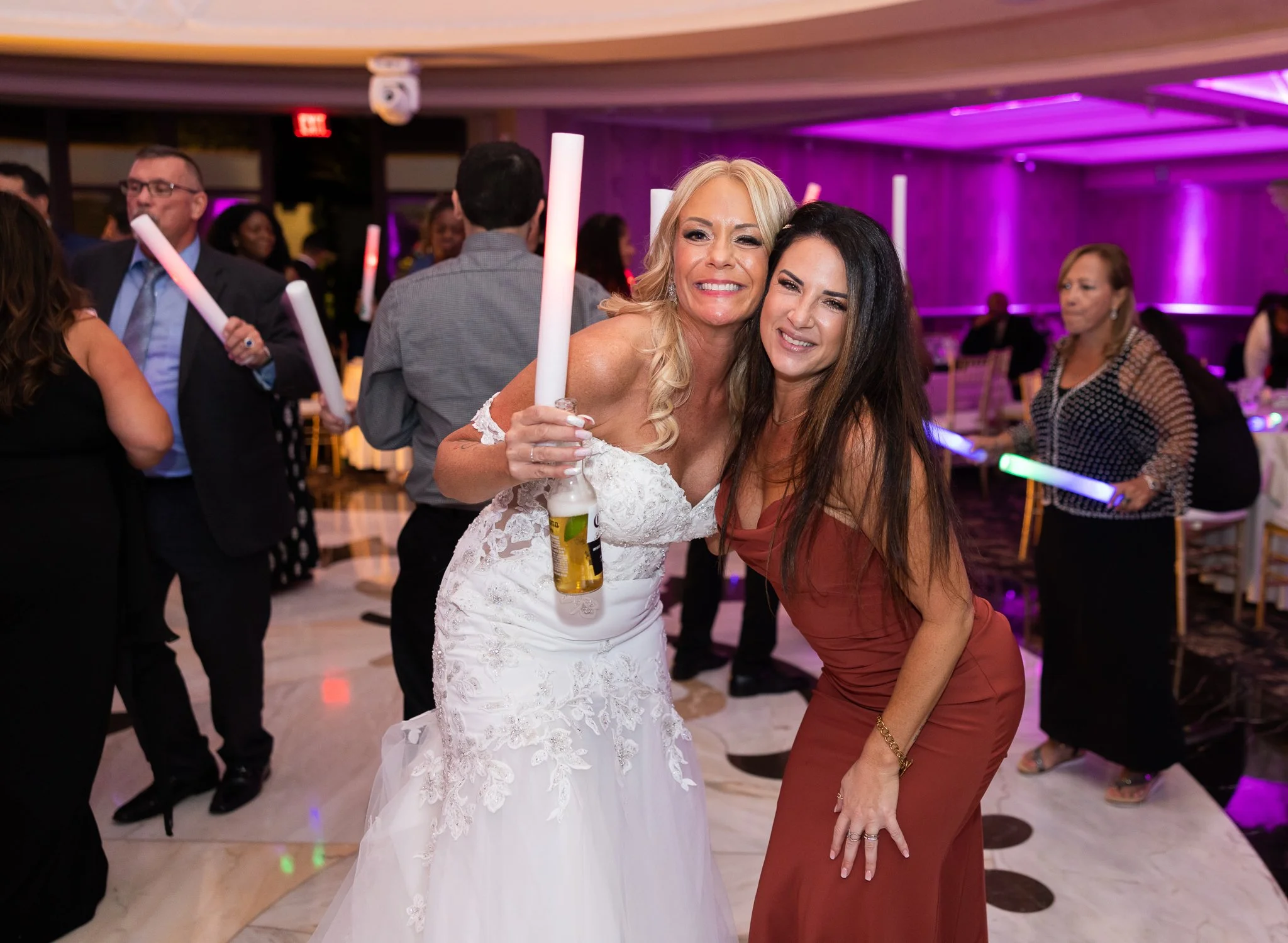 Two women, one in a white wedding dress and the other in a brown dress, smiling and embracing at a celebration party with purple lighting in the background.