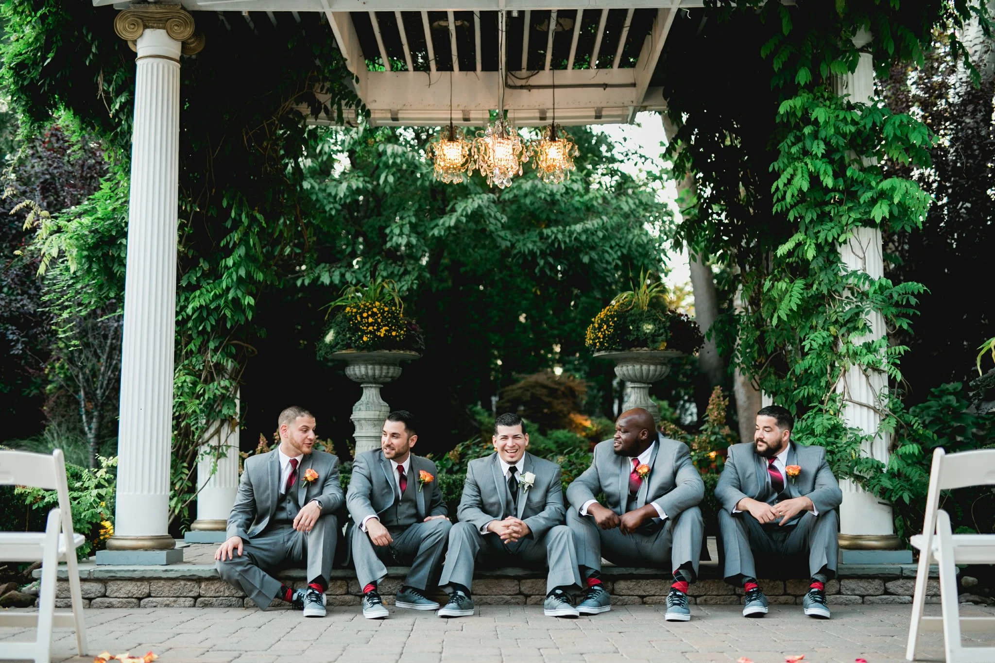 Group of five men in gray suits with orange boutonnieres, sitting on a stone step beneath a white pergola with green ivy, two large planters with yellow flowers, and a chandelier hanging above, in a lush garden setting.