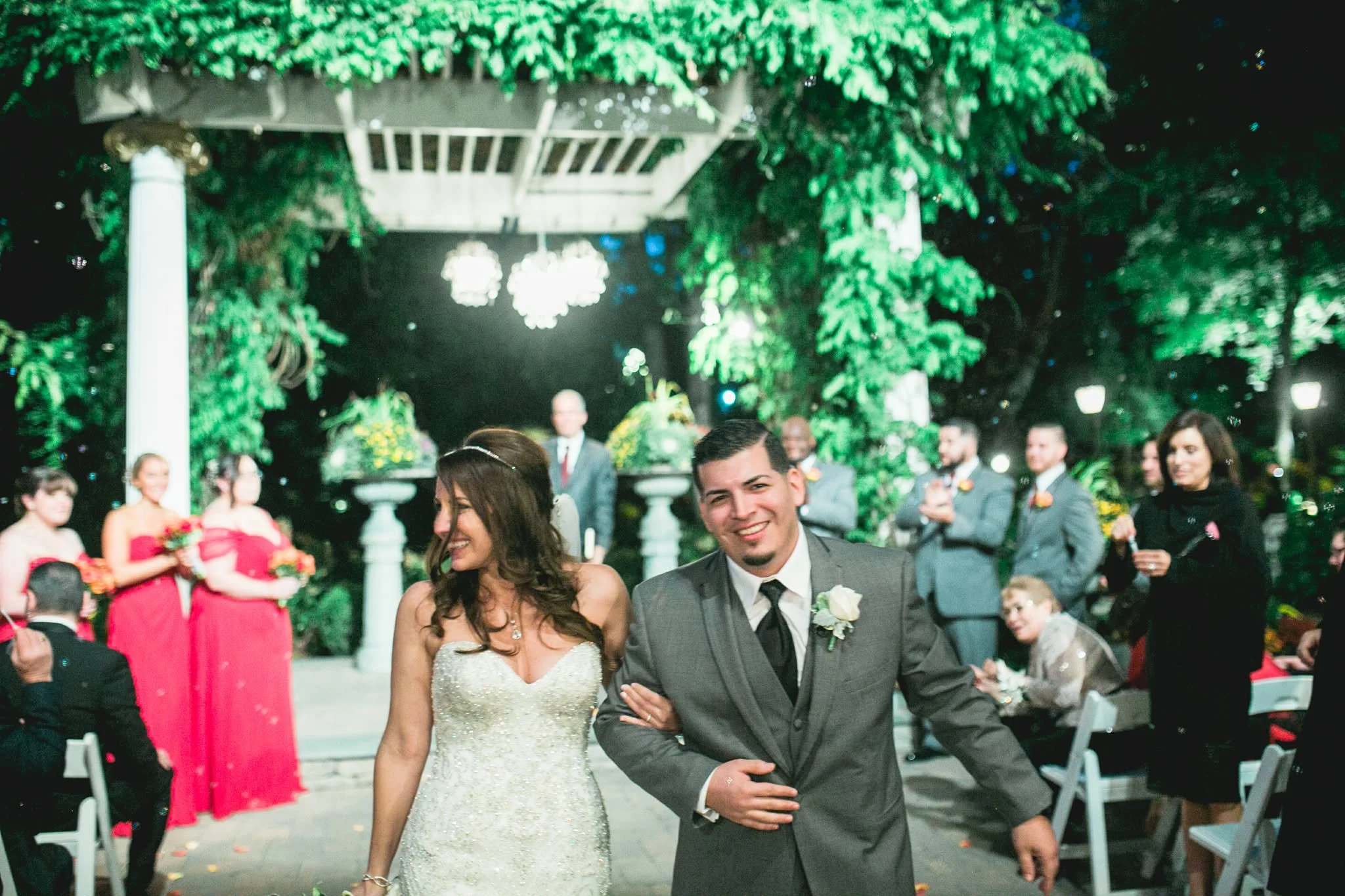 A bride and groom walking together at their wedding reception. The bride is wearing a white strapless wedding gown, and the groom is wearing a gray suit with a black tie. They are smiling and holding hands, with wedding guests in the background under