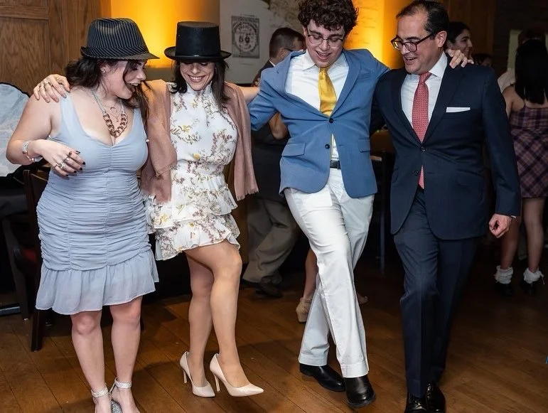 Four people dancing in a line at a party, smiling and holding each other's arms.
