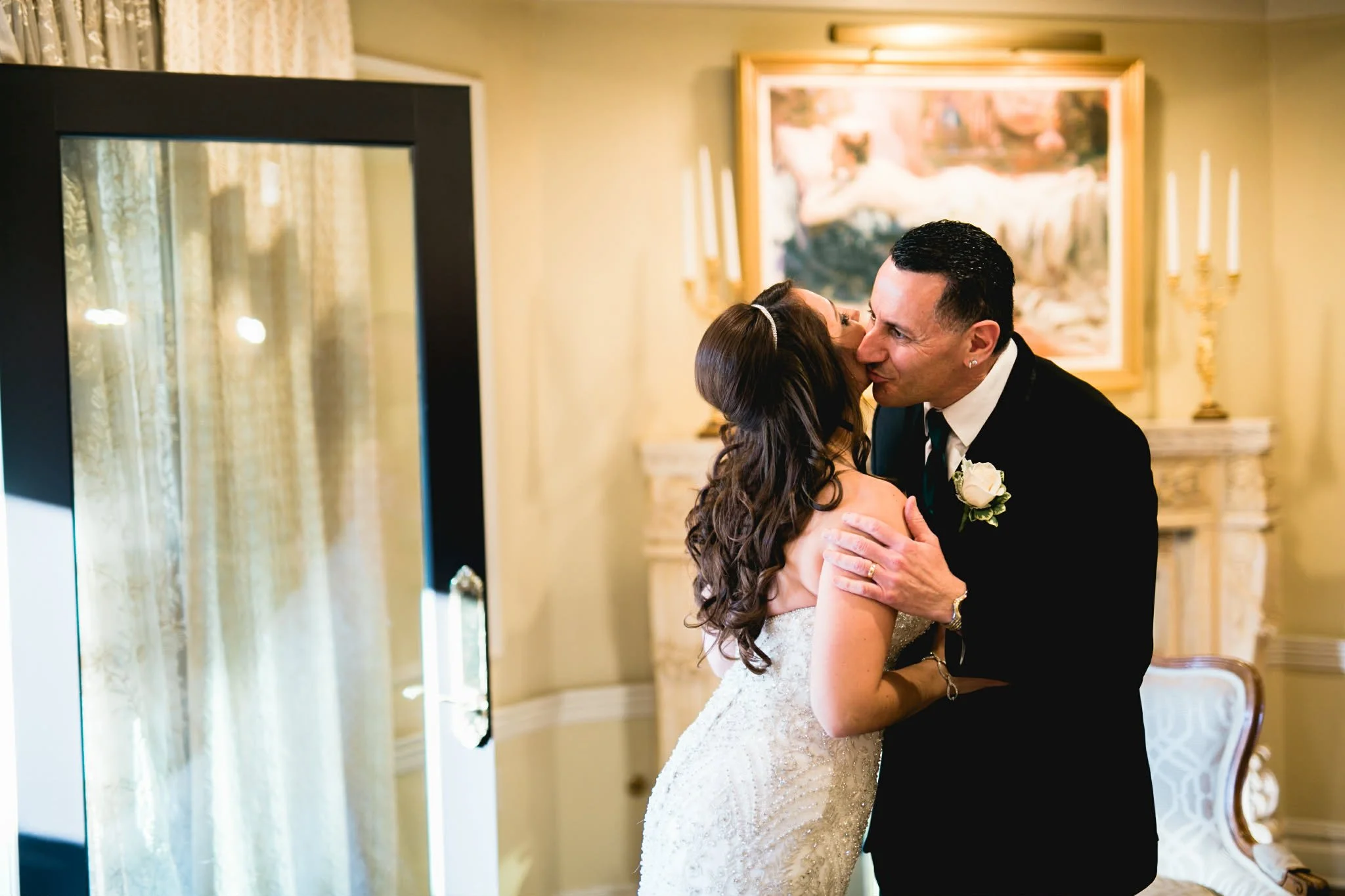 A bride and groom sharing a kiss indoors, with cream-colored walls, a framed painting, and decorative candles in the background.