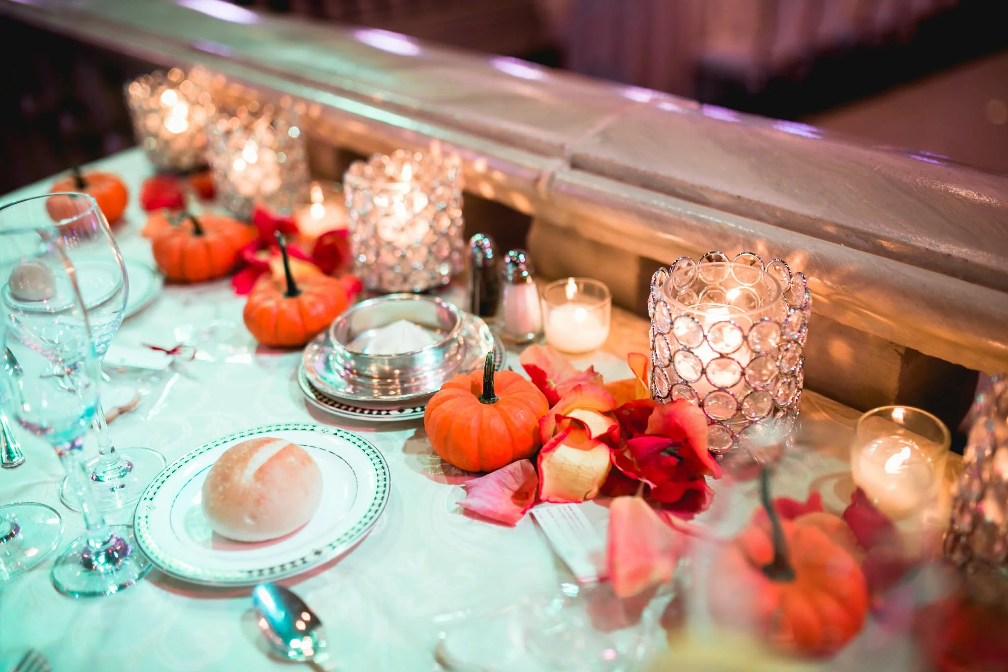 Decorated dining table with small pumpkins, rose petals, and candle holders with lit candles at an elegant event.