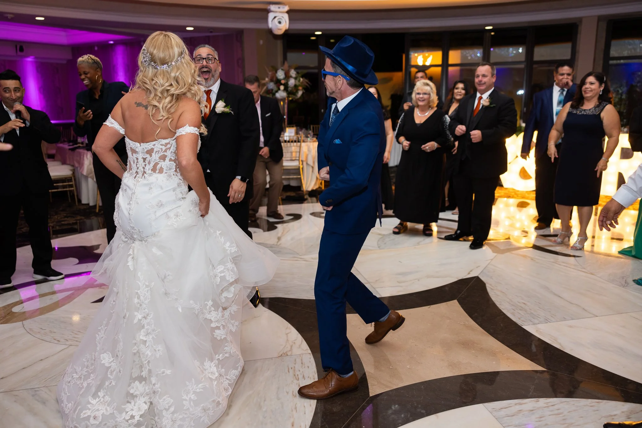 A wedding reception scene with a bride in a white lace wedding gown dancing with a man in a blue suit, with guests watching and smiling in the background, and colorful lighting in the venue.