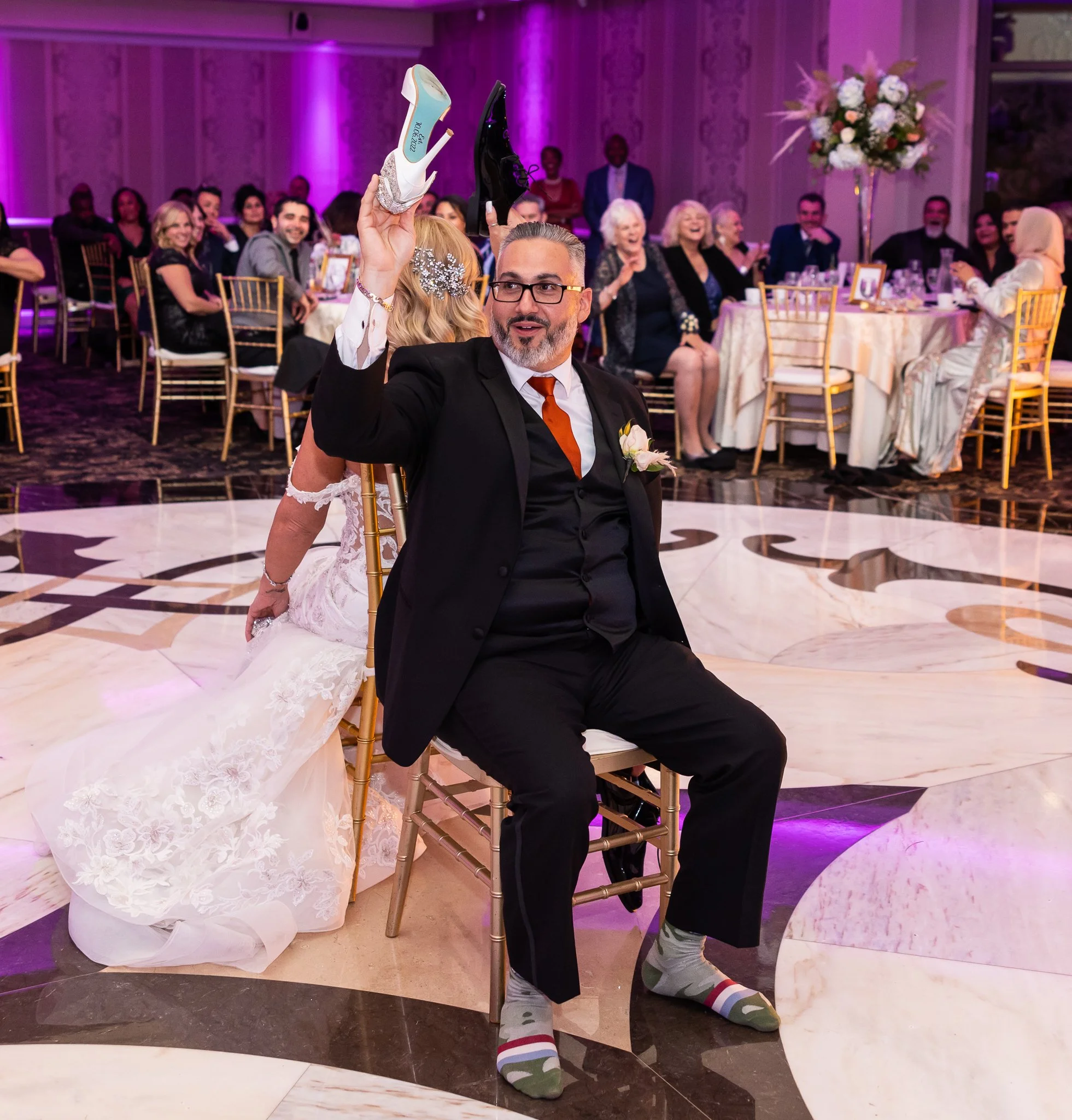 At a wedding reception, a man in a tuxedo with a boutonniere on his jacket sits on a chair holding up a shoe, while the bride, in a wedding dress, kneels behind him. Guests are seated at round tables in the background, with some clapping and smiling,