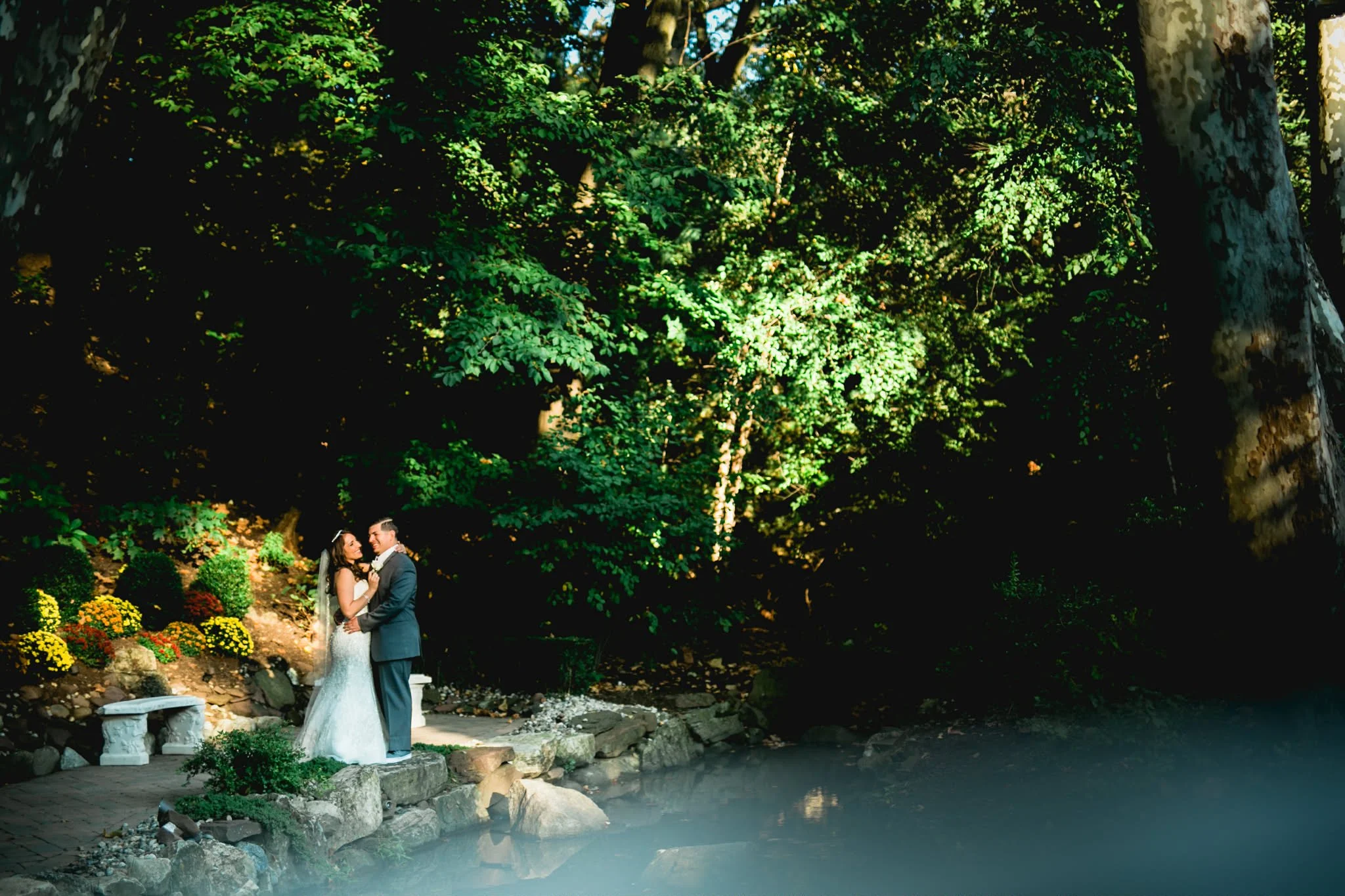 A bride and groom embrace on a stone path near a pond, surrounded by lush green trees and flowering plants, with sunlight filtering through the leaves.