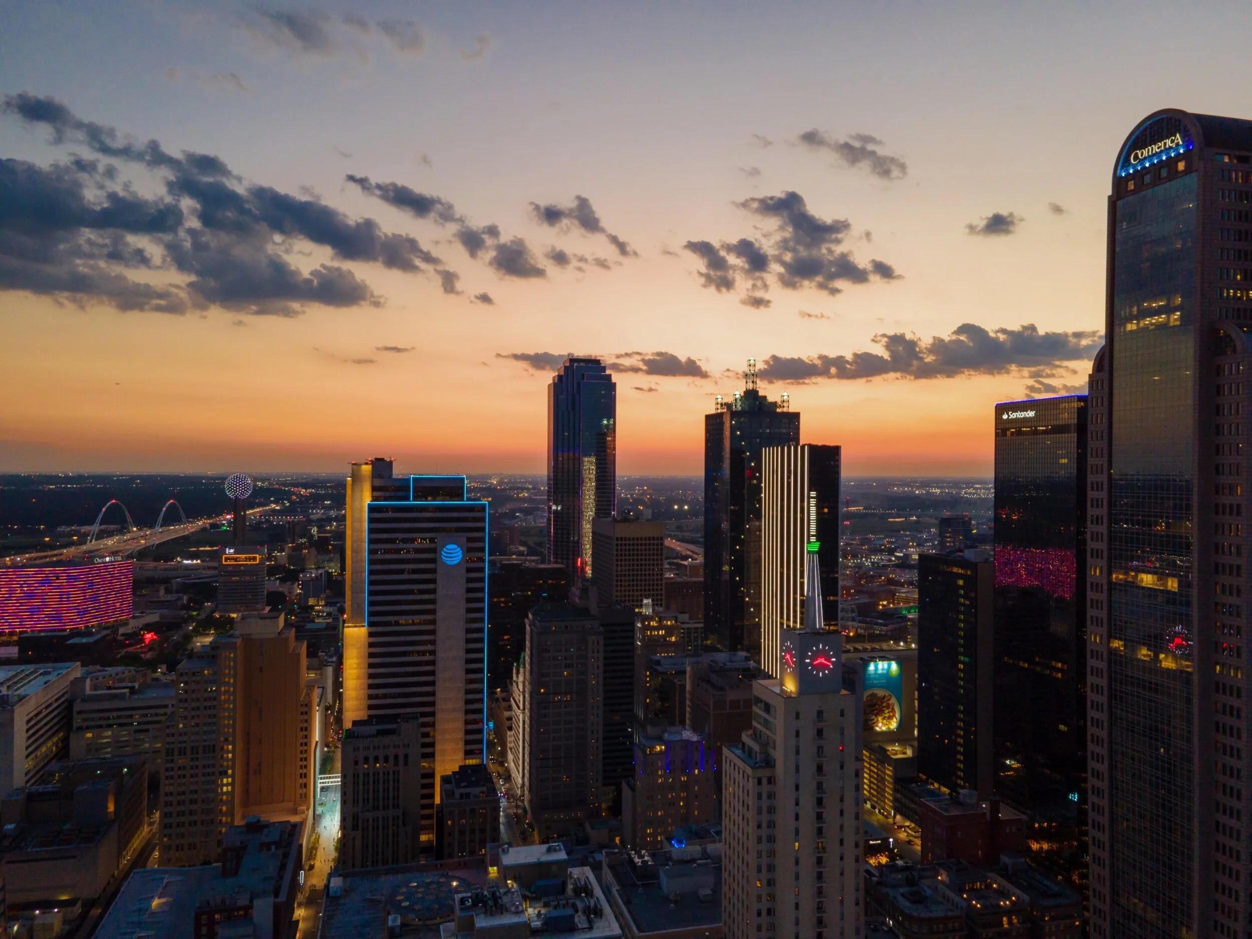 City skyline at sunset with tall skyscrapers, some with illuminated signs, and colorful sky with clouds.