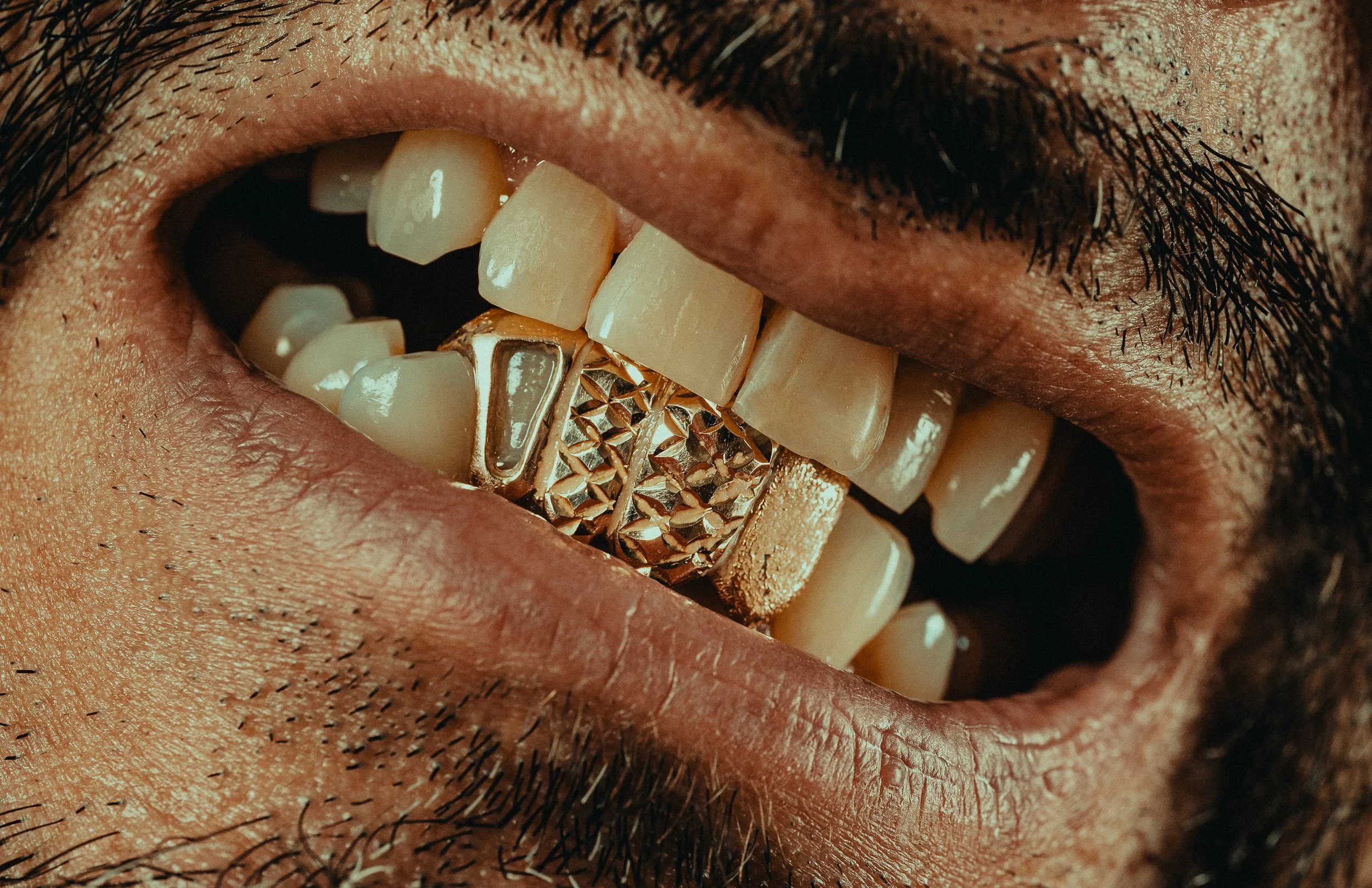 Close-up of a person's mouth with gold grillz on their teeth, slightly open lips, and visible facial hair.