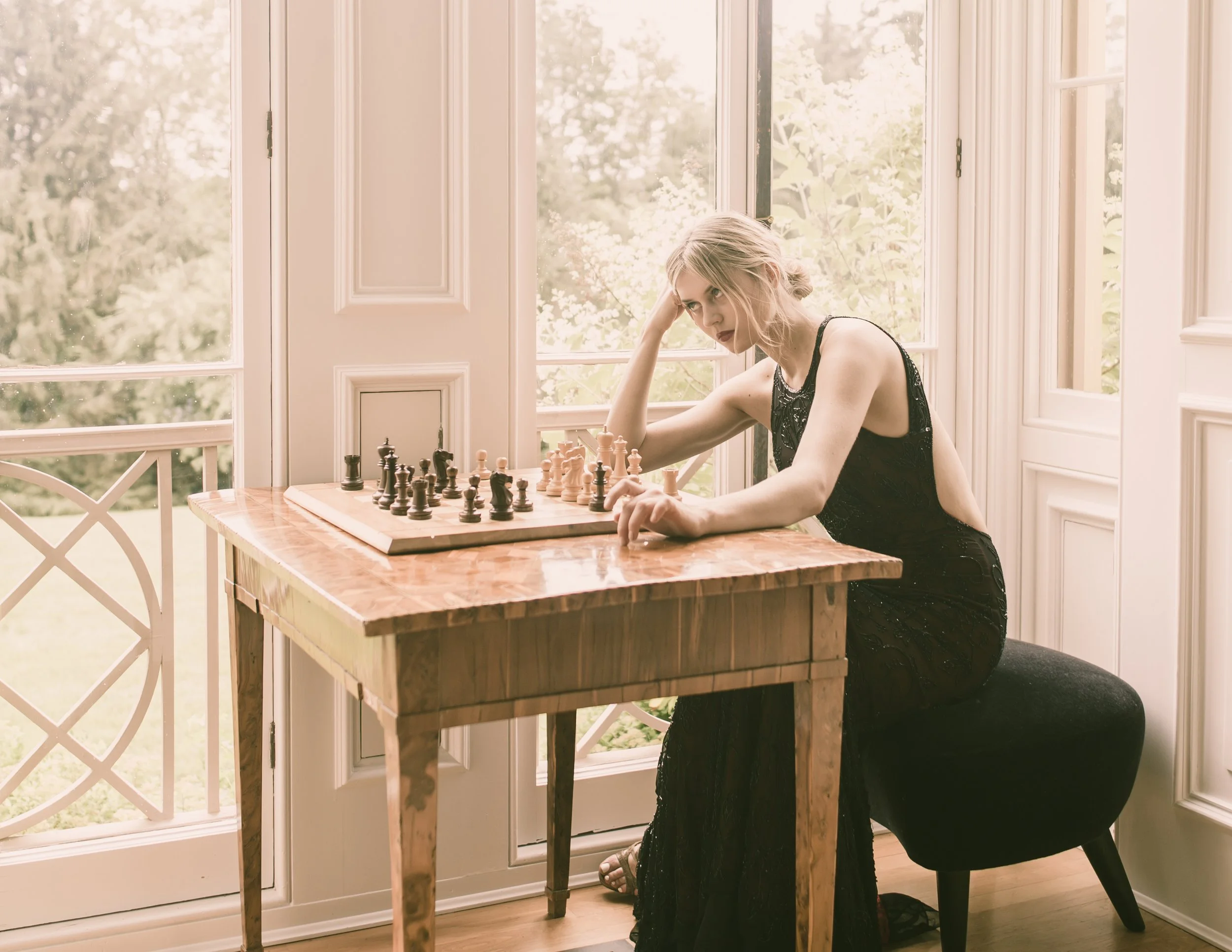 A woman in a black dress is sitting at a wooden table playing chess, looking focused, with a large window showing greenery outside in the background.