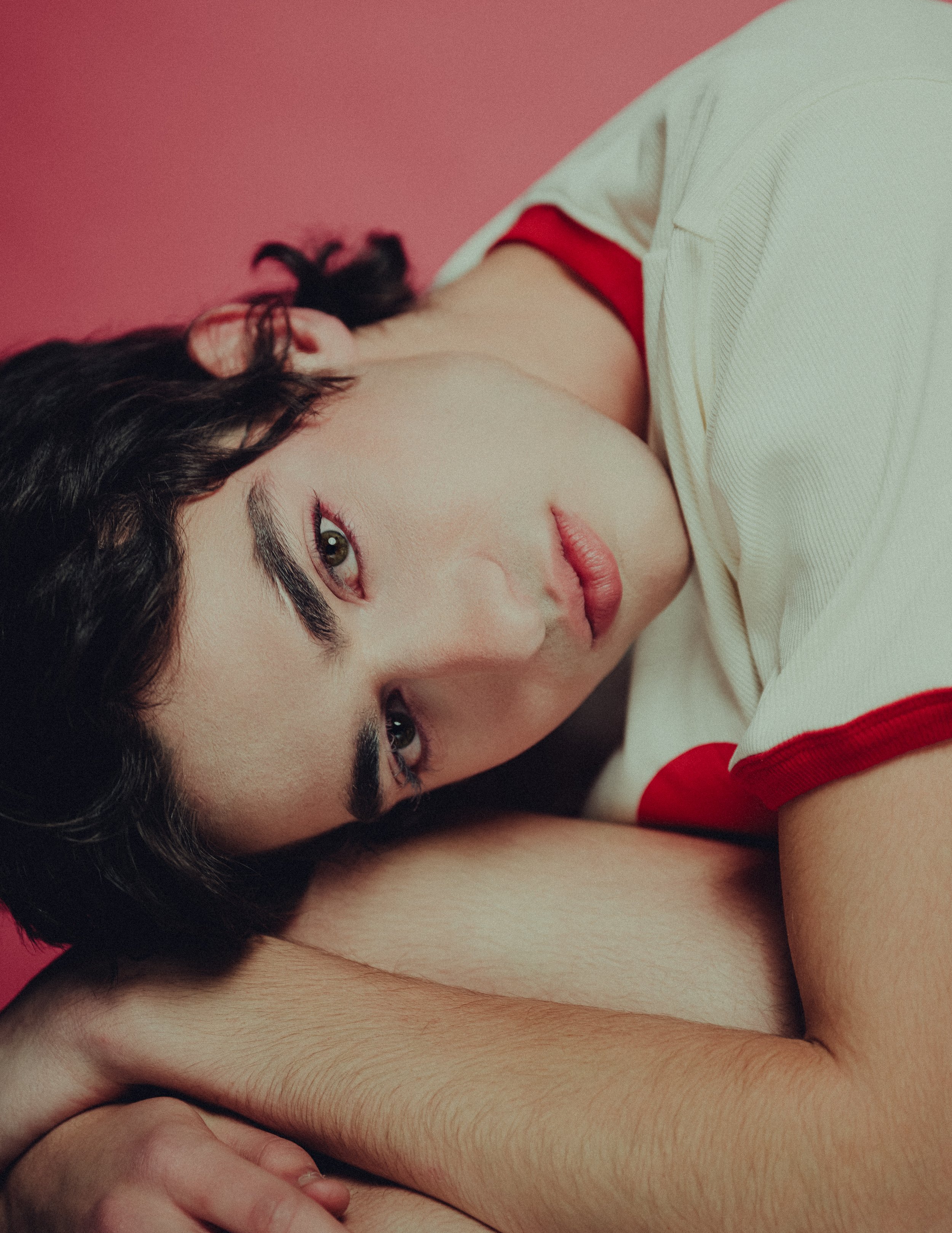 A young woman with dark hair, lying down with her head resting on her knees, looking directly at the camera with a soft expression, against a red background.