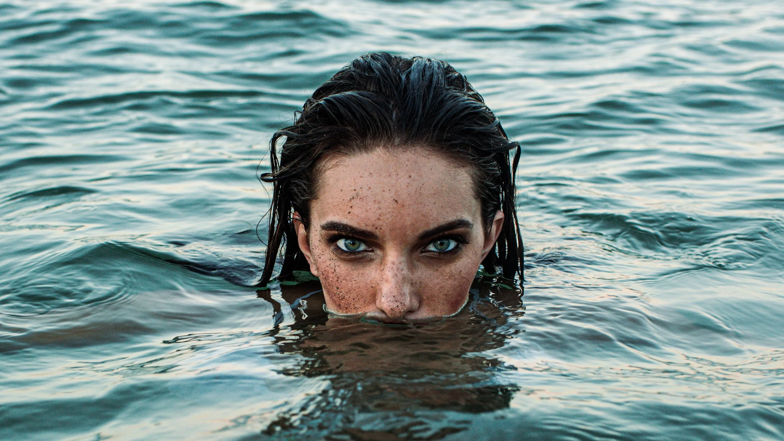 A woman with wet dark hair and blue eyes partially submerged in water, looking directly at the camera.