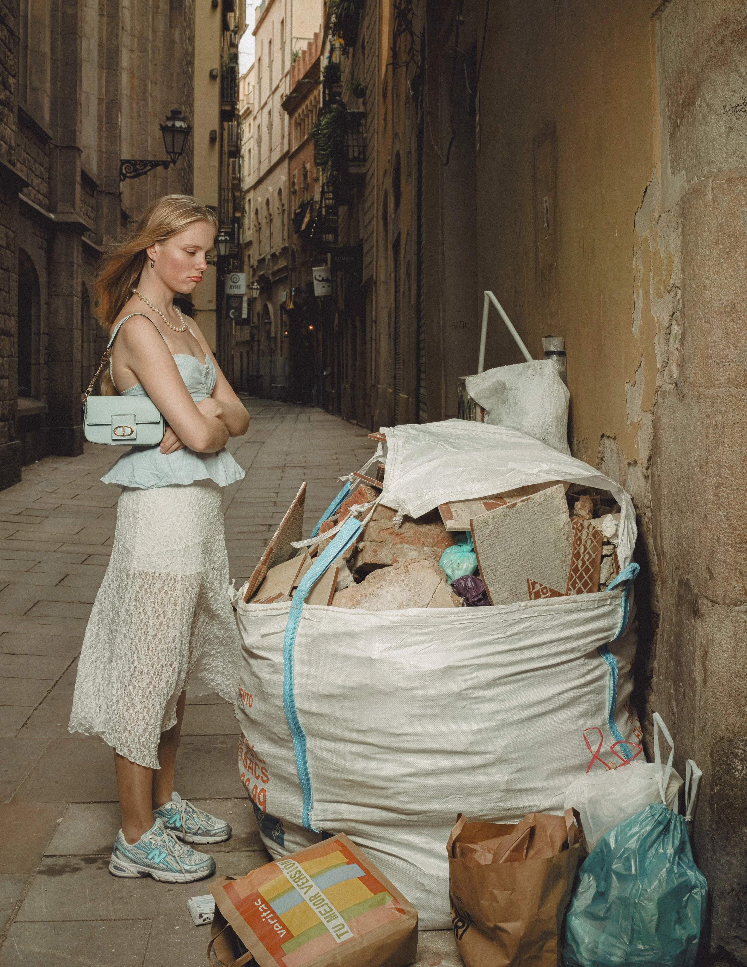 A woman with blonde hair, wearing a light green top, white lace skirt, and teal sneakers, stands with her arms crossed in front of a large pile of trash and debris in a narrow city street.