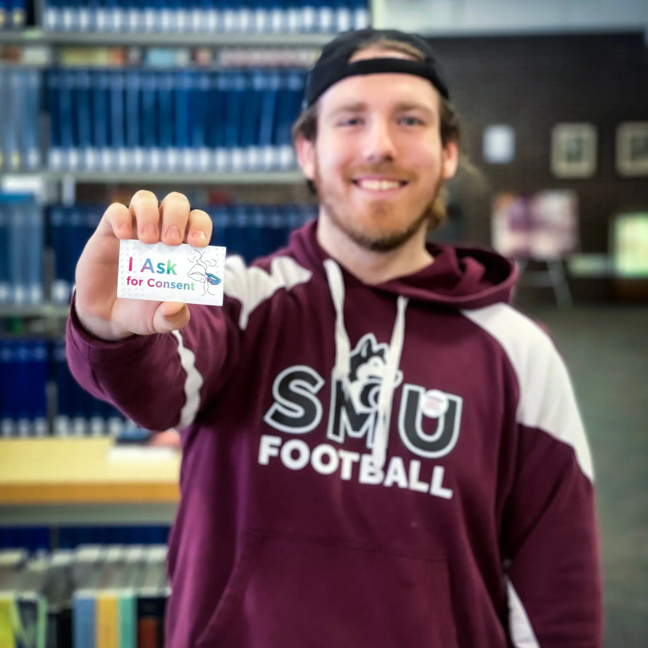 A man in a SMU Football sweater holds a paper that says I ask for consent