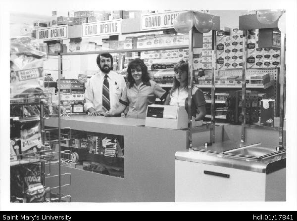  Earl Surette and two female employees stand behind the counter of the Mini Market. 'Grand Opening' signs are all around the store 
