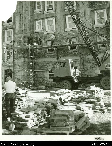  Photograph of north wing of McNally Building, having it exterior stone cladding installed. There are four men on the scaffold, and a figure standing in the left foreground amidst stacks of stone.  &nbsp; 