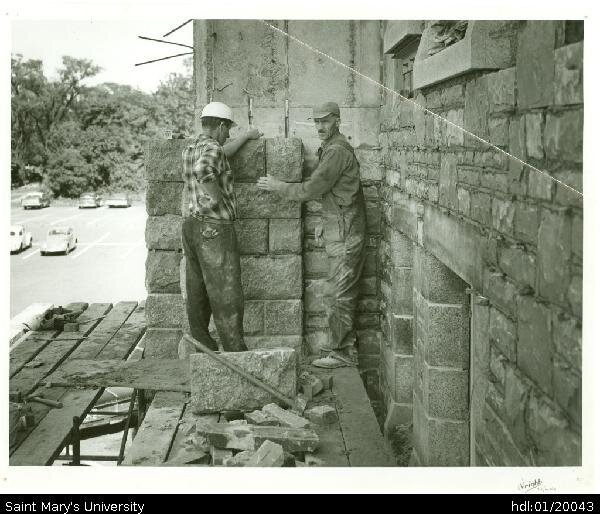  Two men adding stone cladding to exterior wall of McNally north wing. The picture appears to be facing east, towards the current site of the Student Centre. Also shown is a parking lot with five cars. Accompanying note reads: 'McNally building under