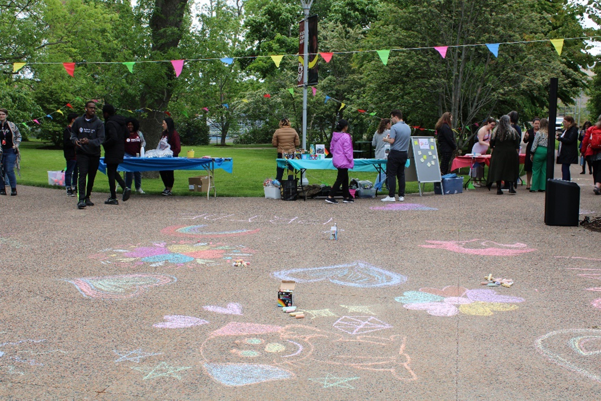  Saint Mary's Quad featuring several Pride activity tables along edge of grass and colorful chalk drawings on cobblestone path.   