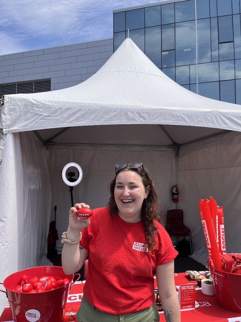  Emma, who has light skin and long dark hair, holds a mini football as she stands smiling in front of the Keep It Social promotional booth 