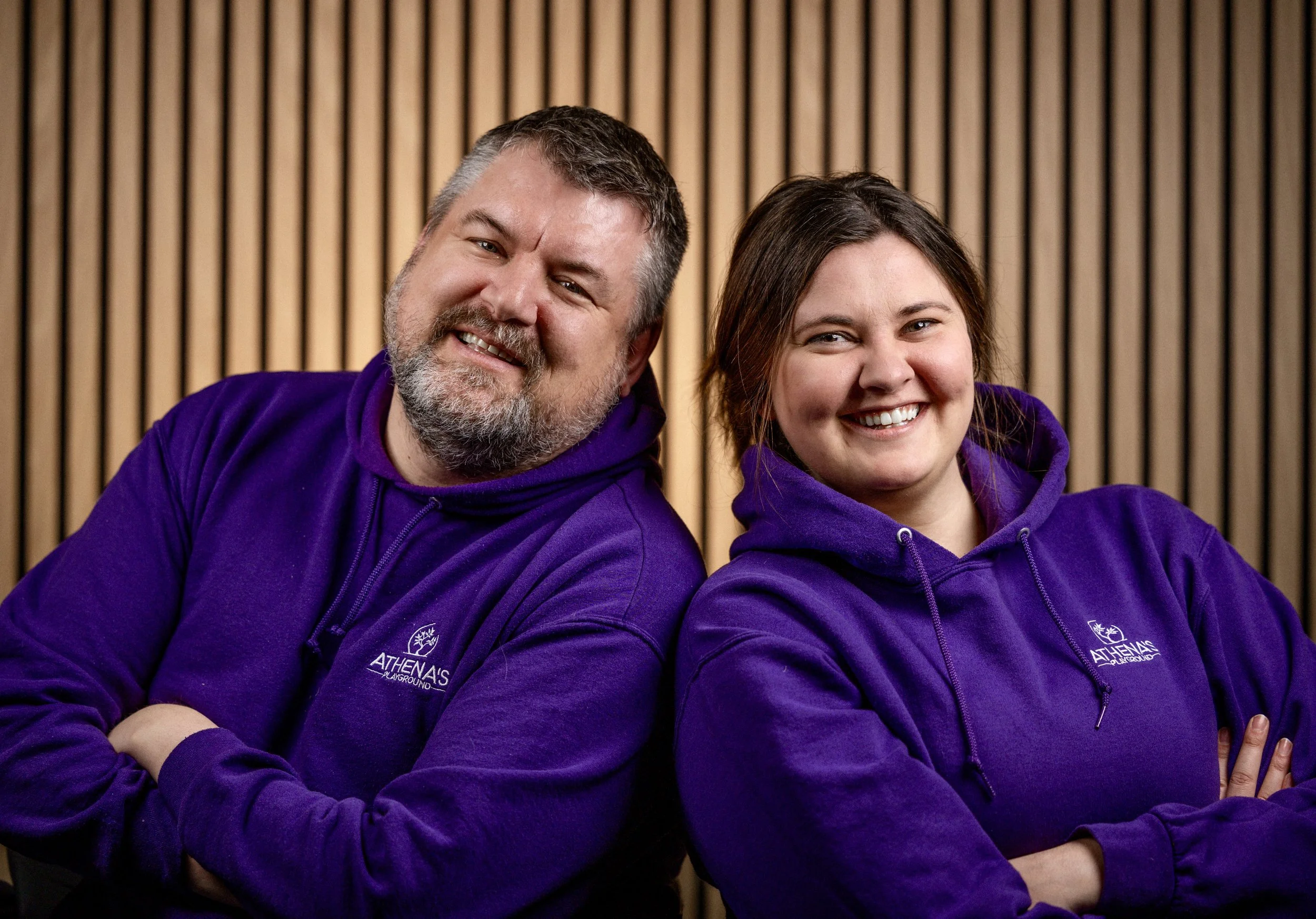 Michael and Meredith stand with their arms crossed while smiling. They wear purple branded Athena's Playground hoodies