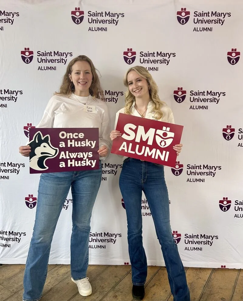Maddie and a friend hold SMU alumni signs and stand in front of a SMU alumni backdrop