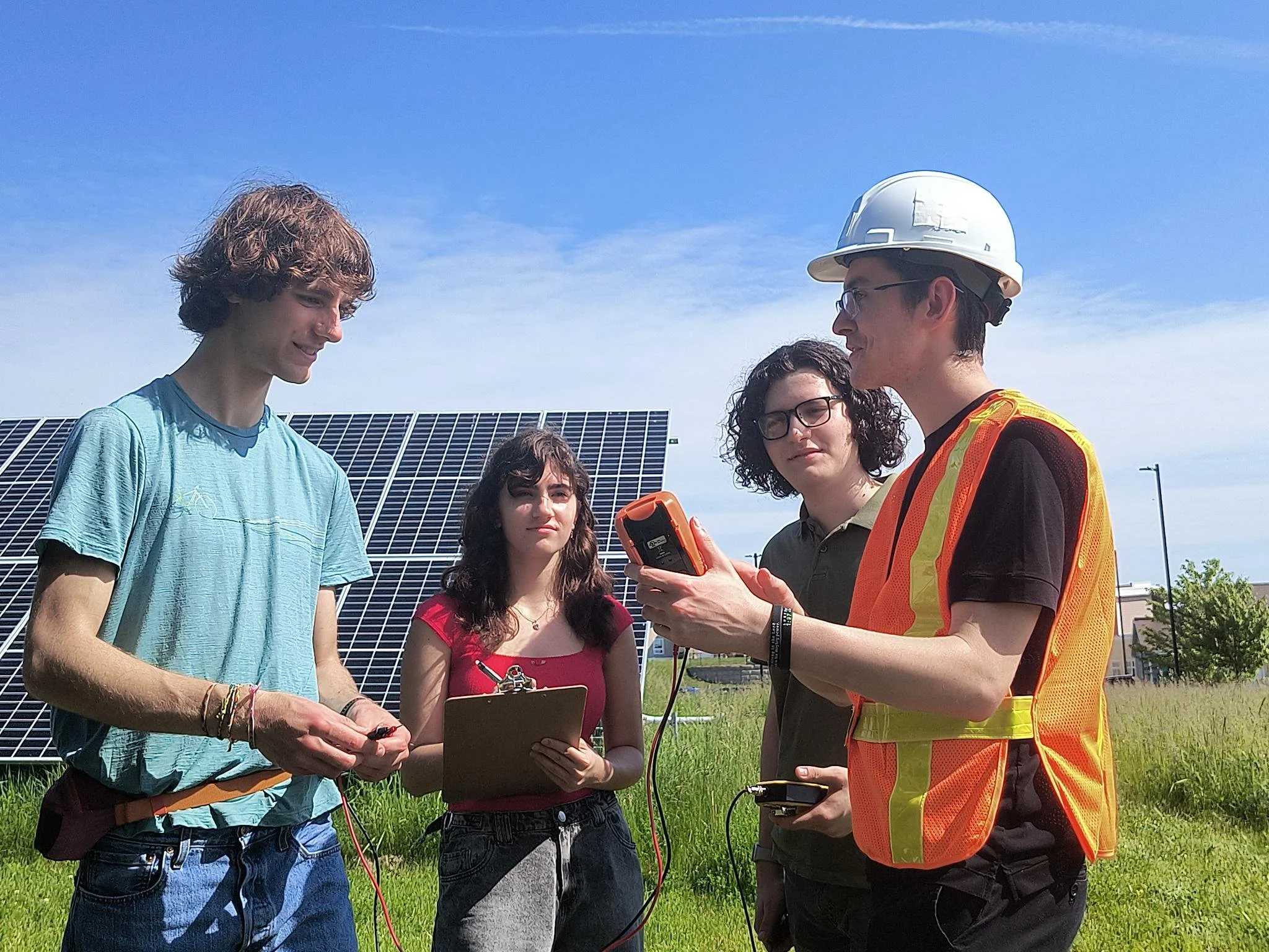 Four people stand outdoors near an industrial solar panel. Students take notes while the guide in safety gear gives a demonstration