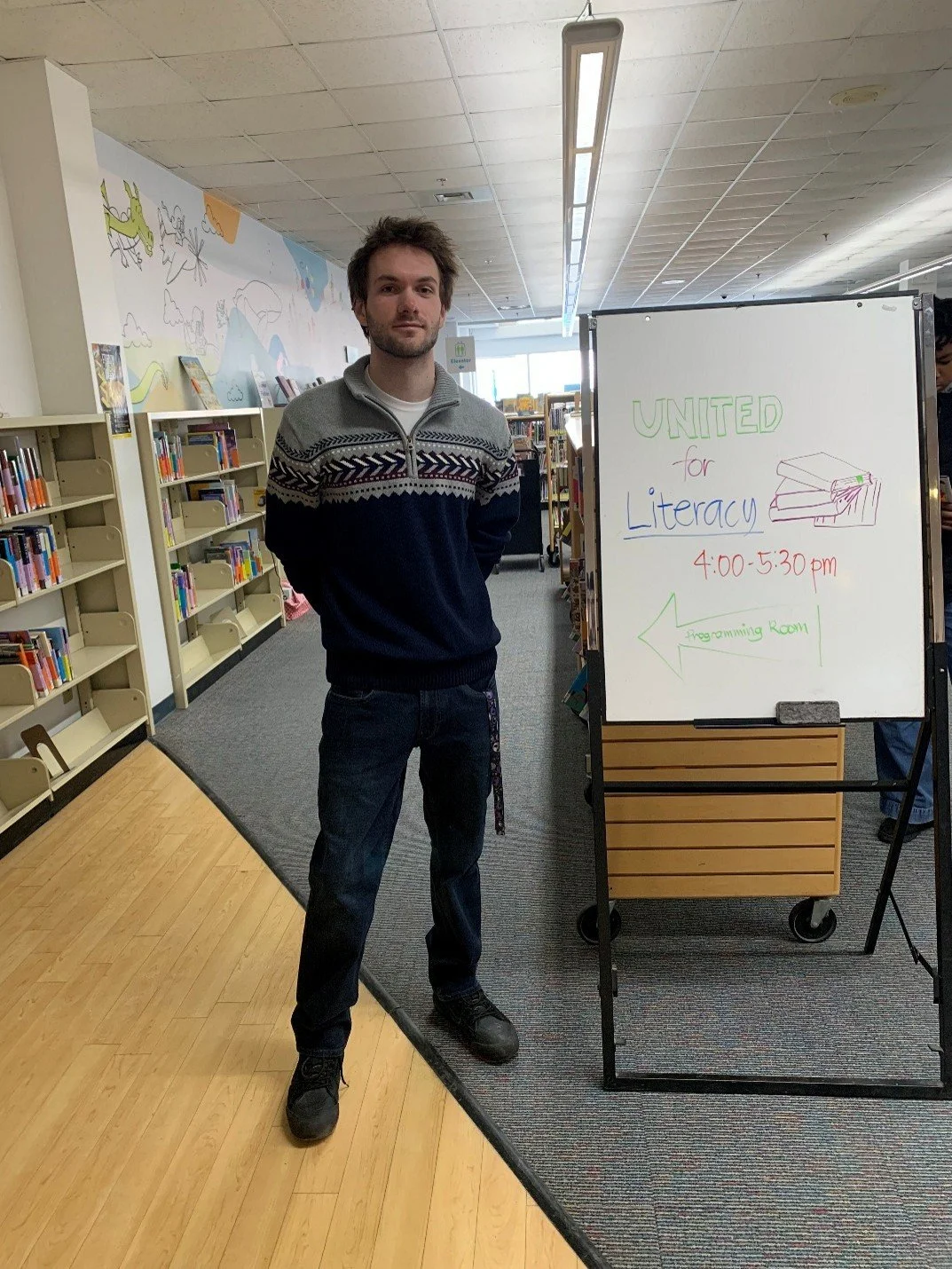 Matthew stands inside a library next to a sign which reads ' United for Literacy'
