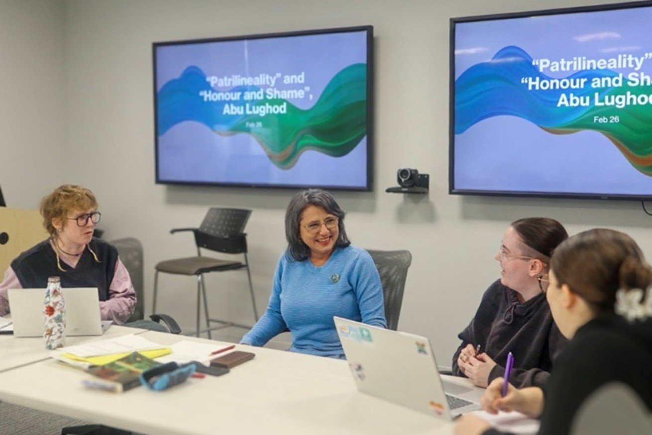 Dr. Takseva sits at a table surrounded by smiling students, a presentation shown on screen behind them