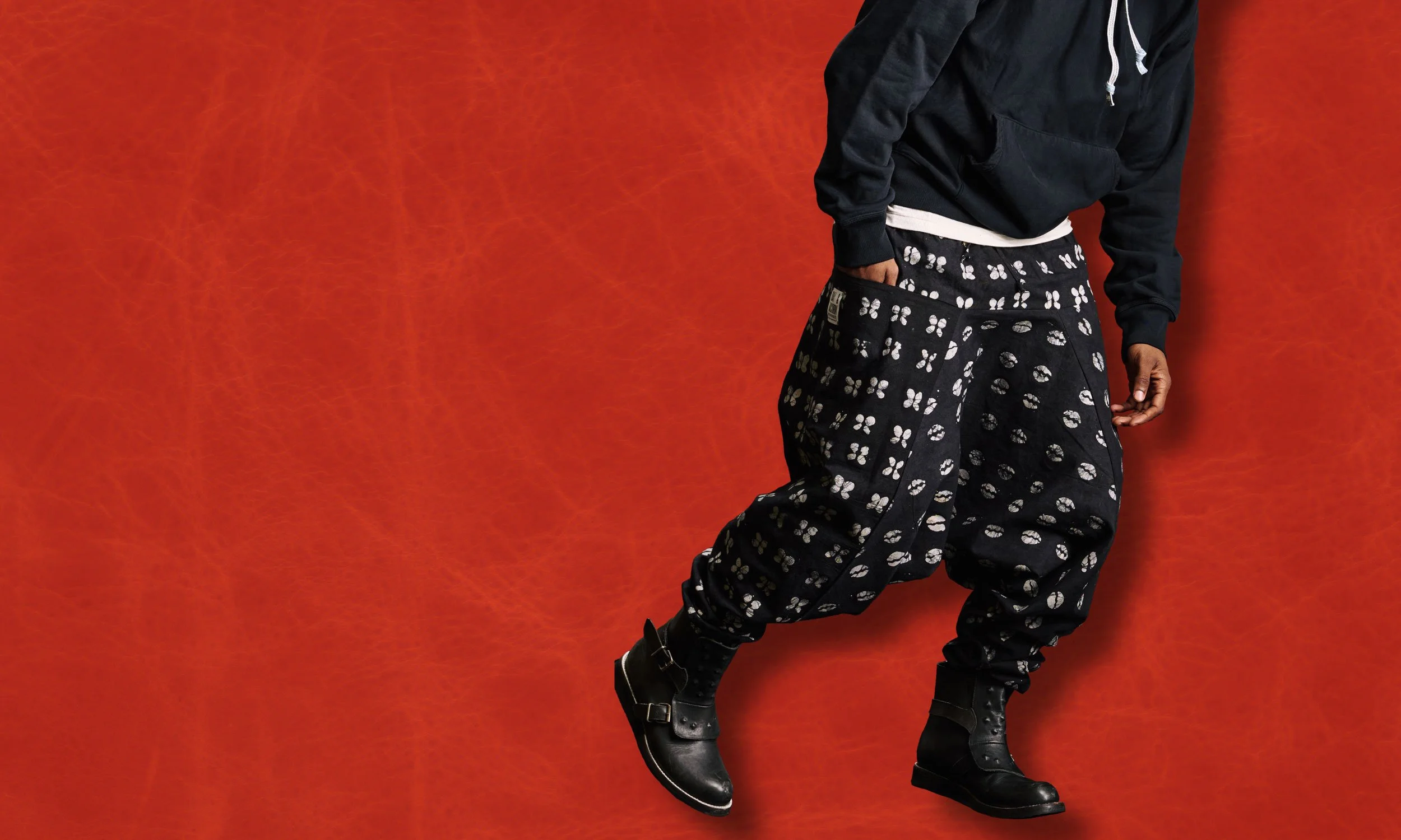 Two men wearing black hoodies and black baggy pants with white patterns, standing against a brown textured background.