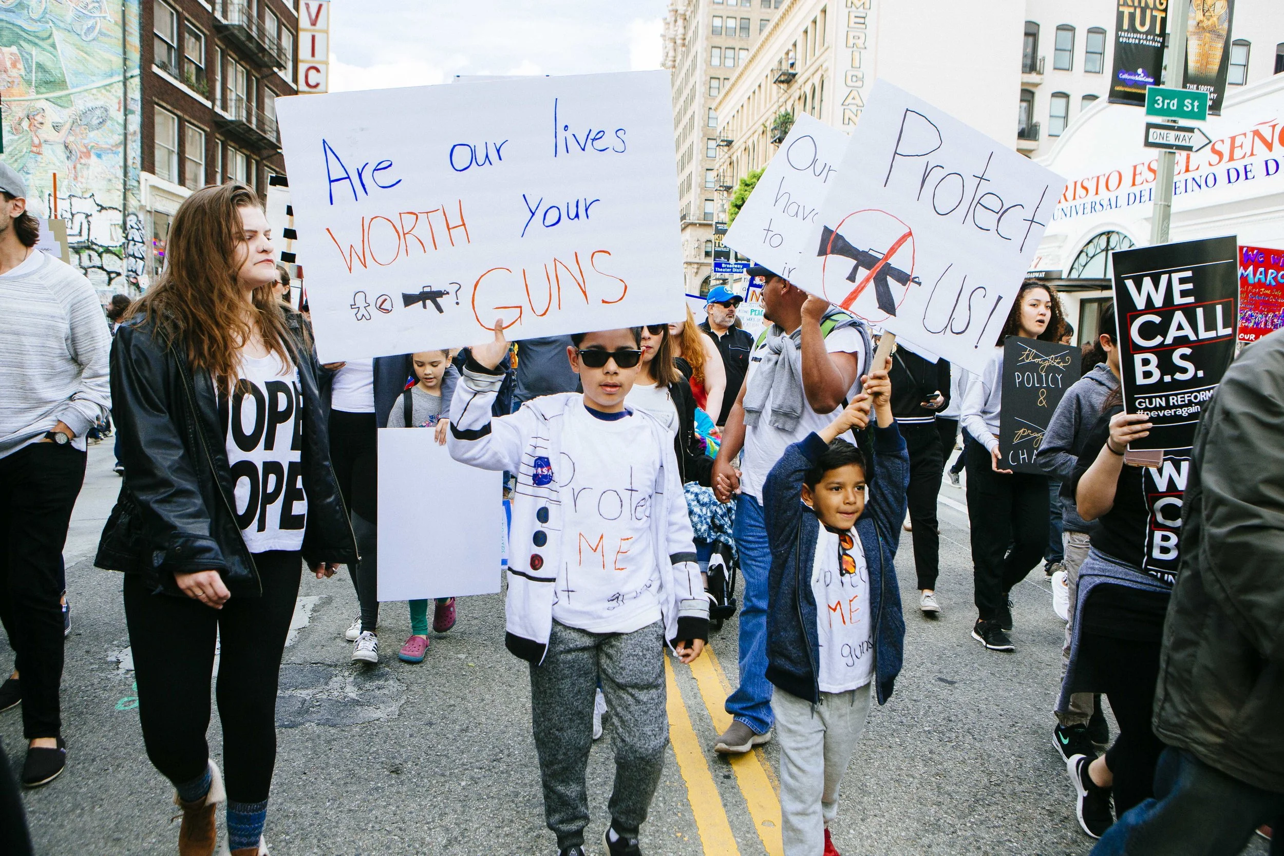 [Image Description: Two children hold signs in the center of a street. Their signs say "Are our lives worth your guns?" And "Protect Us!" with a crossed out drawing of a gun. 