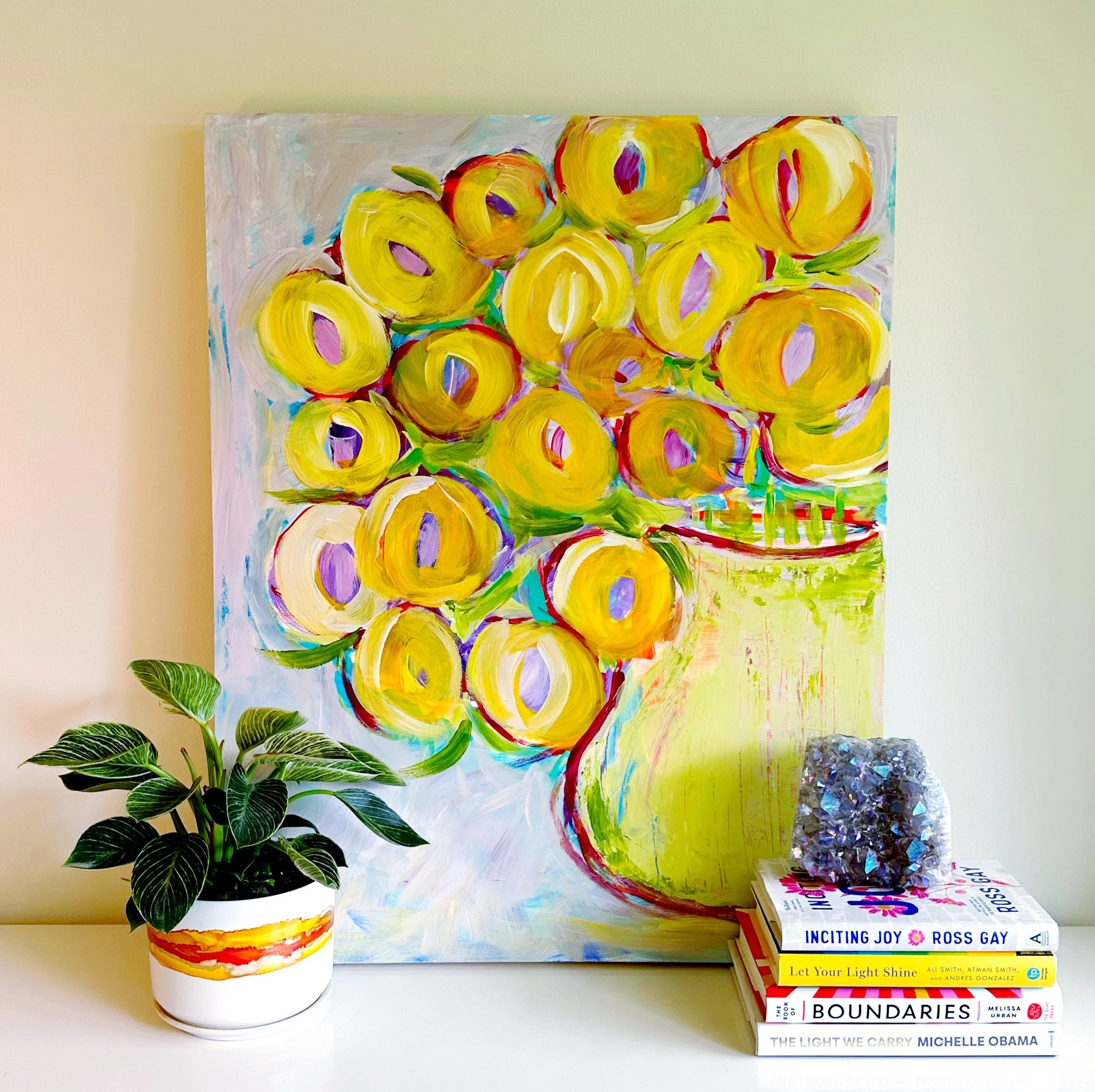 Colorful abstract painting with yellow, orange, purple, and red swirling shapes, plants on the left, and a stack of books on the right.