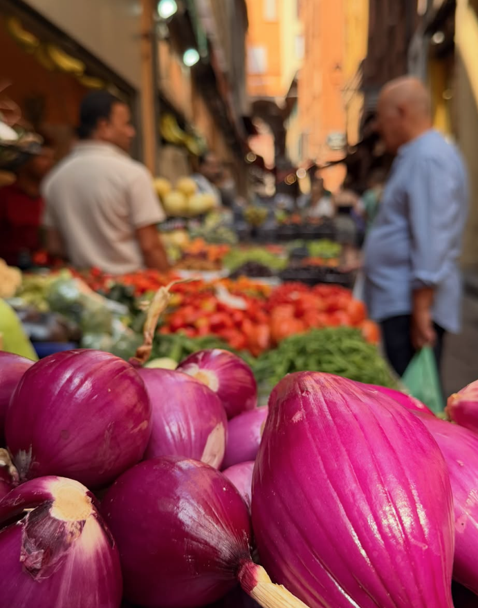 Close-up of pink onions at a market stall with fresh vegetables in the background and market stalls on a narrow street.