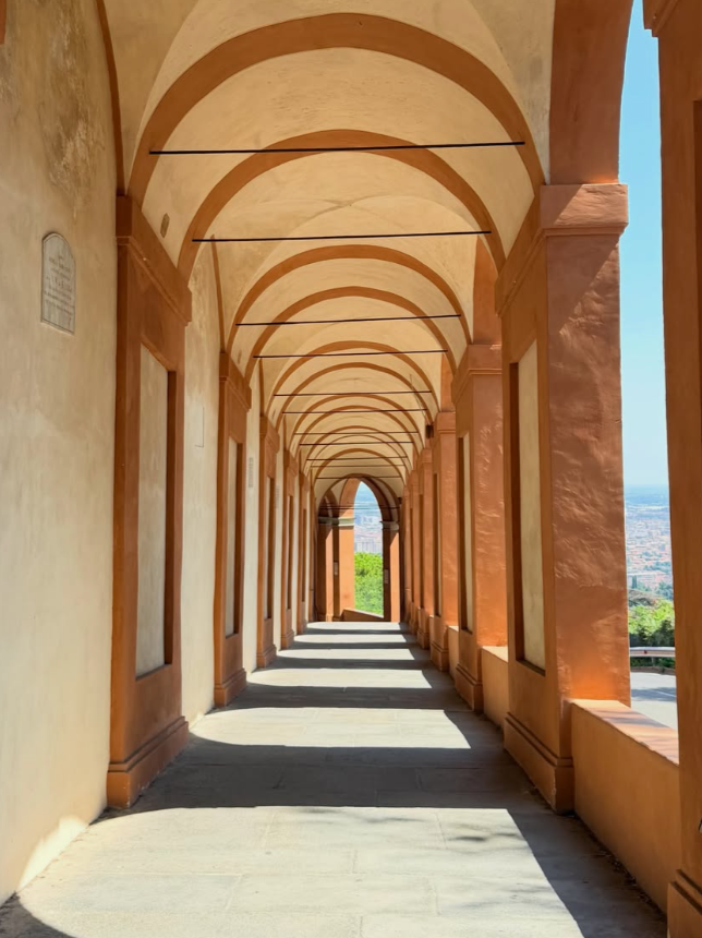 A covered outdoor walkway with a series of arches and columns, overlooking a cityscape and greenery in the distance.