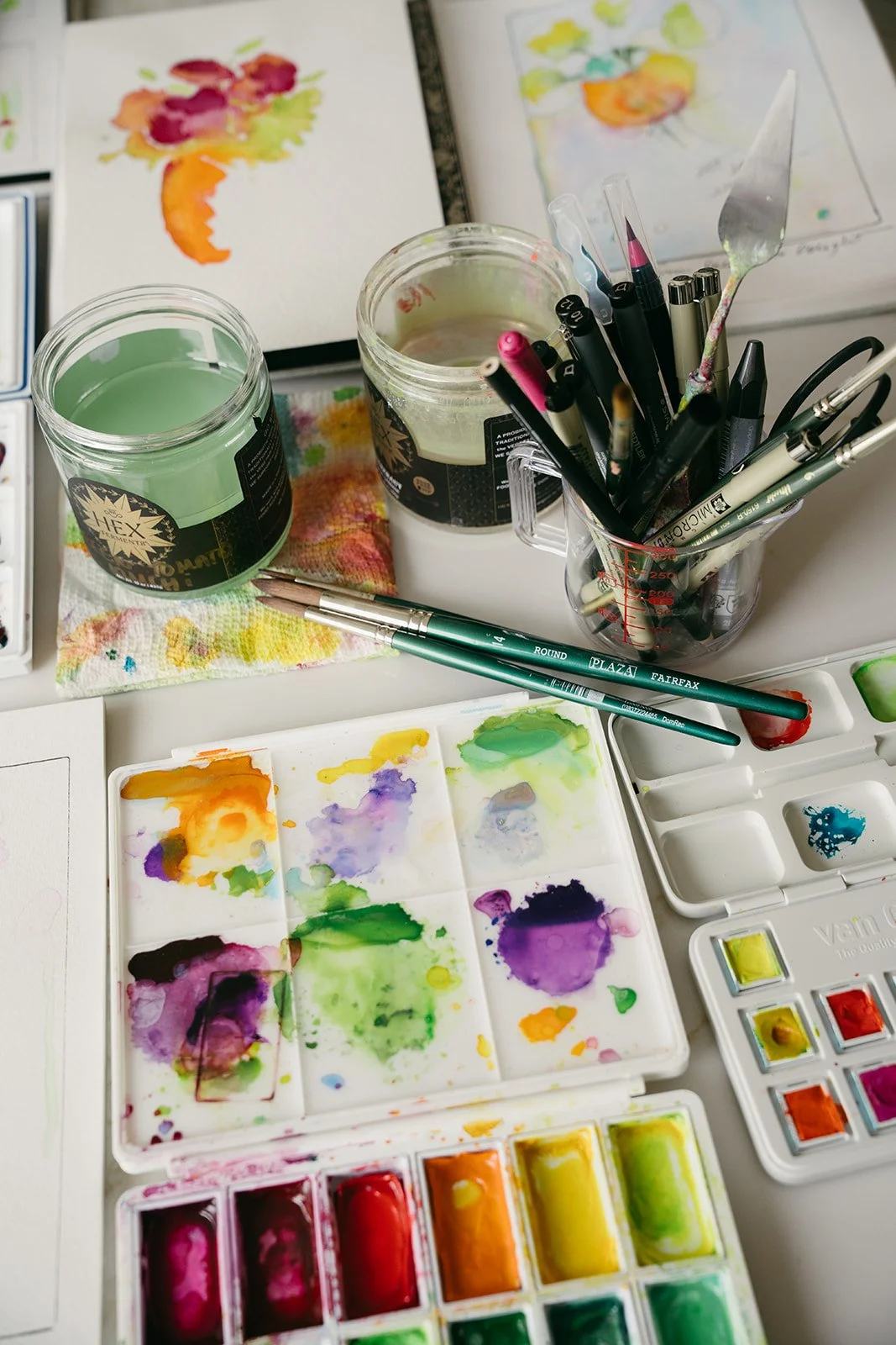 Art supplies including watercolor paints, brushes, pens, and water jars on a workspace with sketches of watercolor paintings.