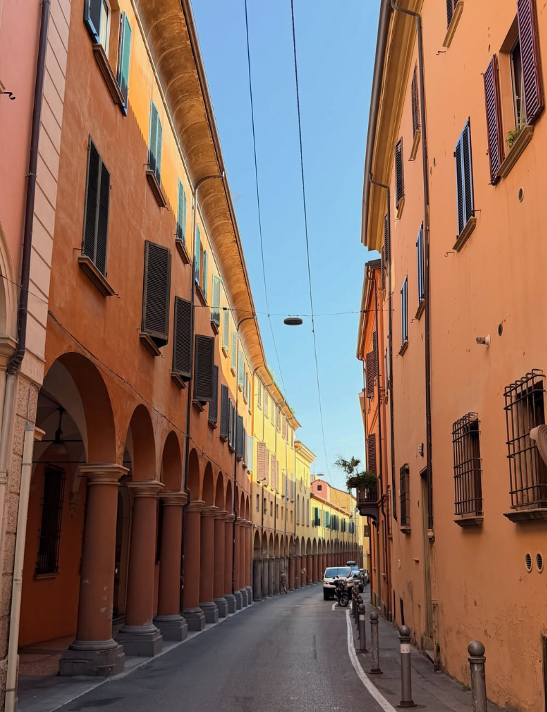 Narrow street lined with colorful buildings on both sides, featuring arched walkways on the left and windows with shutters, with parked cars and street bollards along the curb under a clear blue sky.