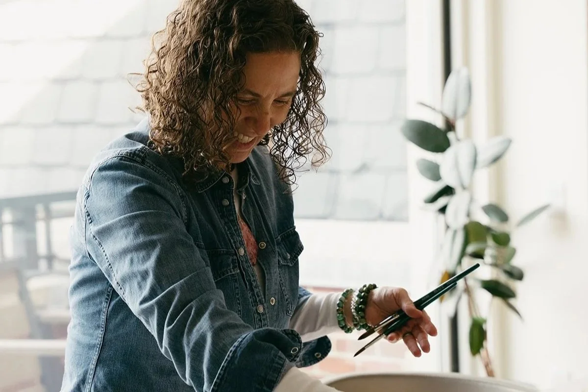 A woman with curly hair wearing a denim jacket holding paintbrushes and smiling inside a bright room with window and potted plant.