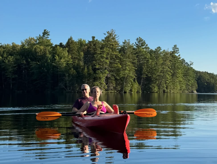 Two people kayaking on a calm lake with a forested shoreline, under a blue sky with some clouds.