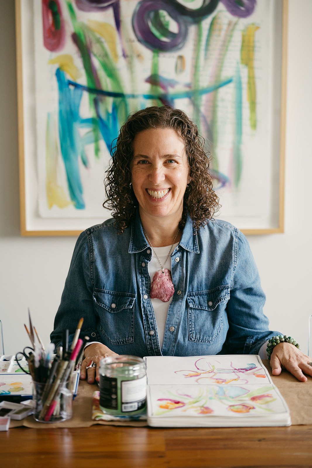 A woman with curly hair smiling and sitting at a table with art supplies and watercolor paintings, with an abstract colorful painting in the background.