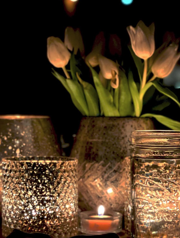 A dark scene with a bouquet of white tulips in a gray pot, surrounded by lit candles in textured glass holders.