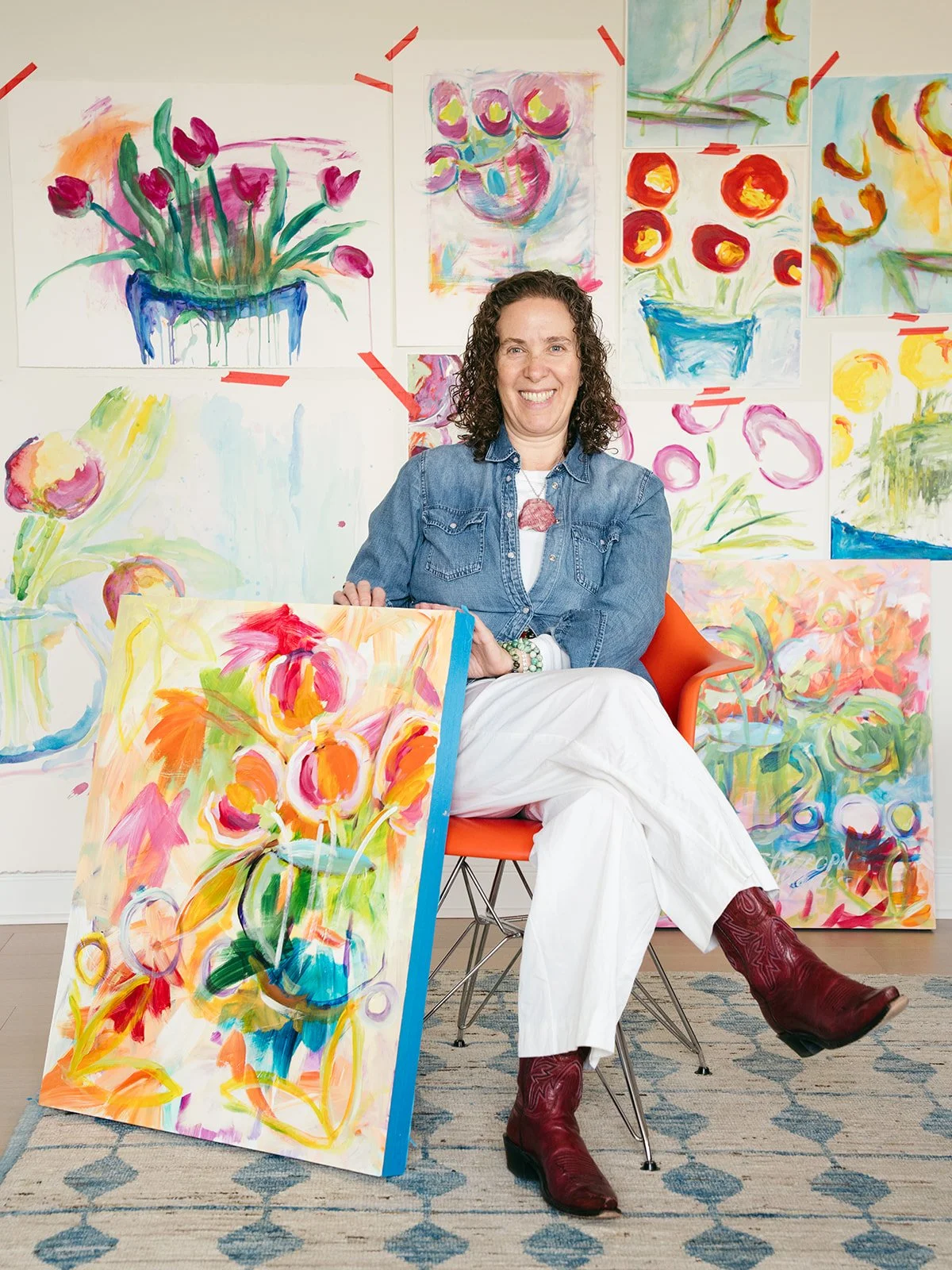 A woman with curly hair sitting on an orange chair surrounded by colorful abstract floral paintings, holding a vibrant painting of flowers. She is smiling and dressed in a denim jacket, white pants, and red boots.