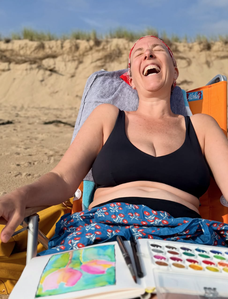 Woman laughing joyfully while sitting outdoors in a beach chair, with painting supplies and a watercolor painting of flowers in front of her.